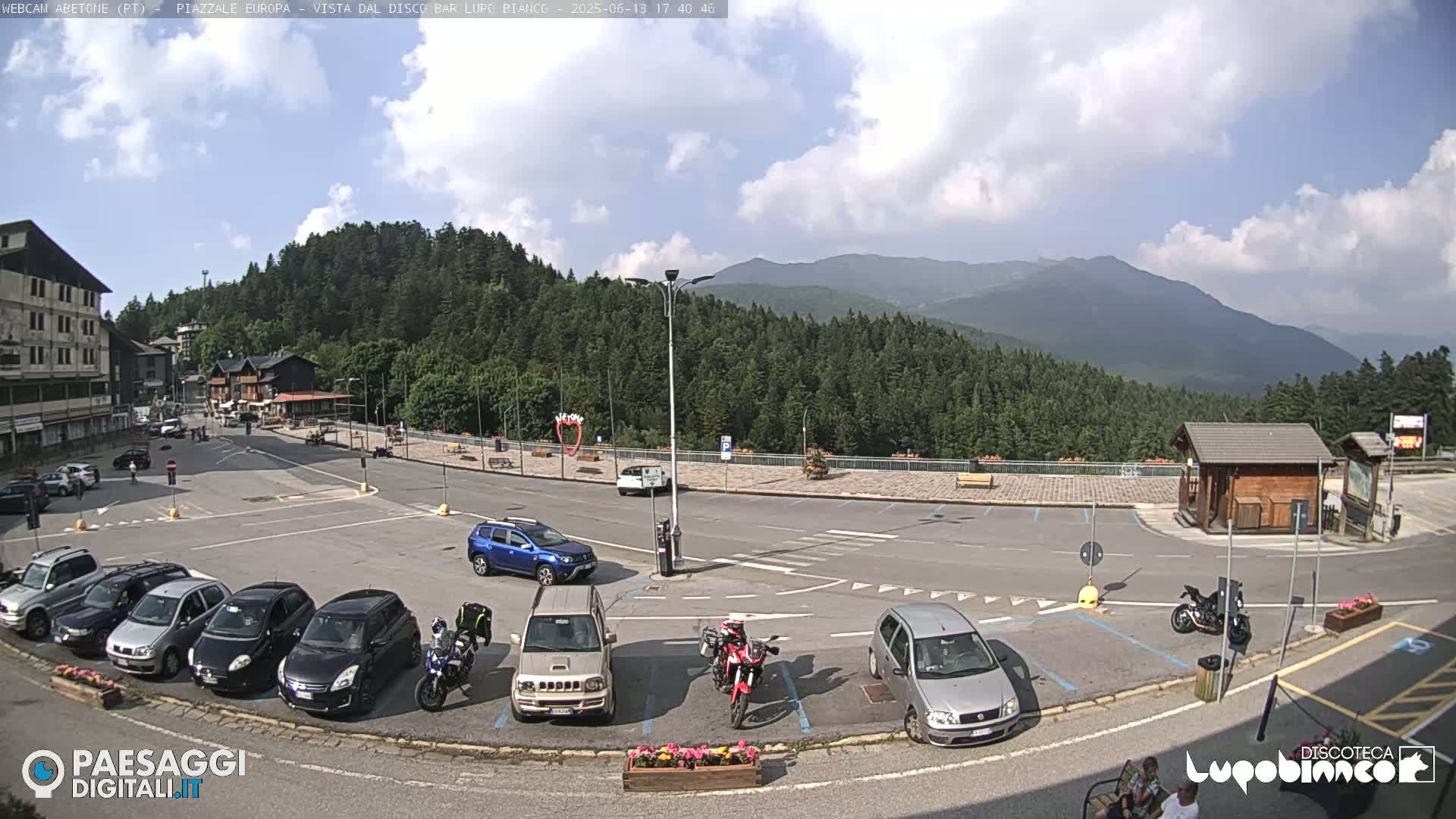 A mostly sunny day shows a mountain view from a parking lot filled with cars and motorcycles, near some buildings and a small wooden structure.