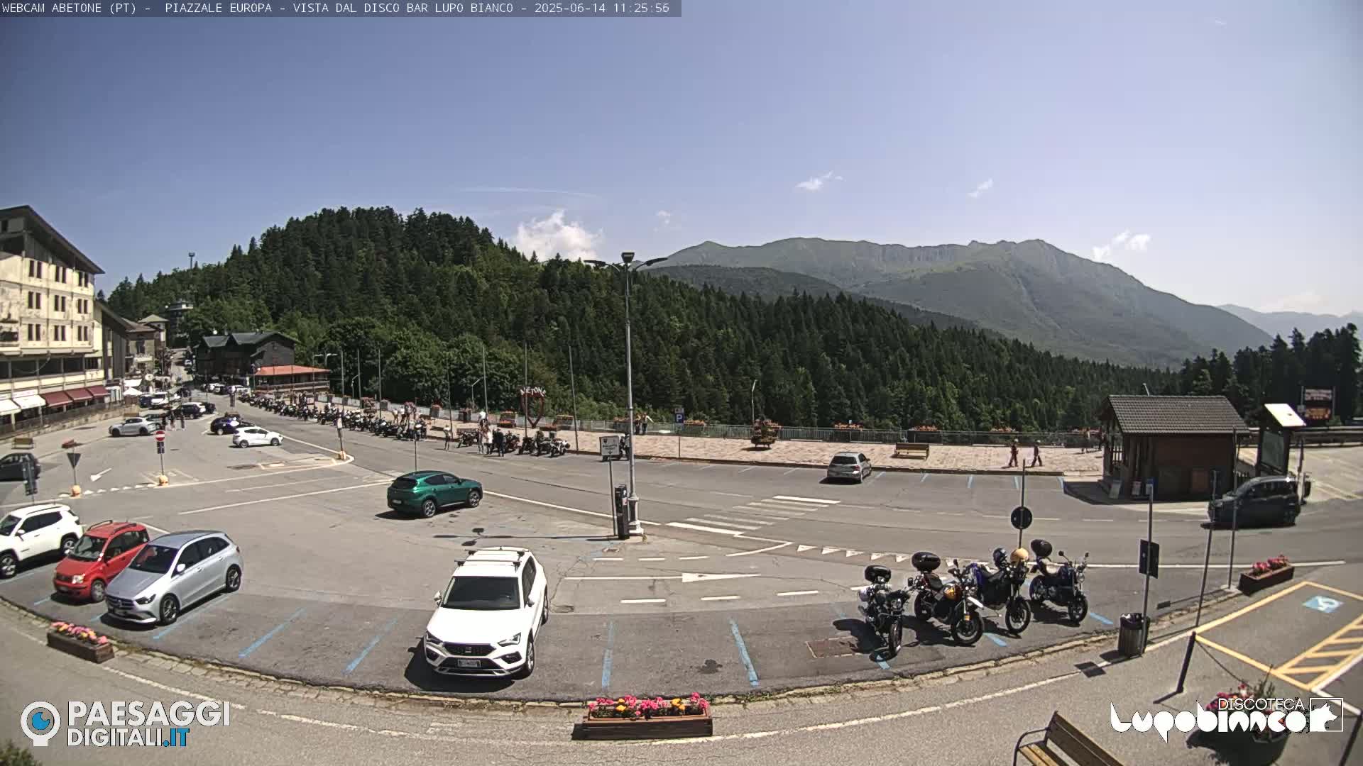 A sunny mountaintop parking lot with cars and motorcycles, overlooked by a building and surrounded by a lush green forest and distant mountains.