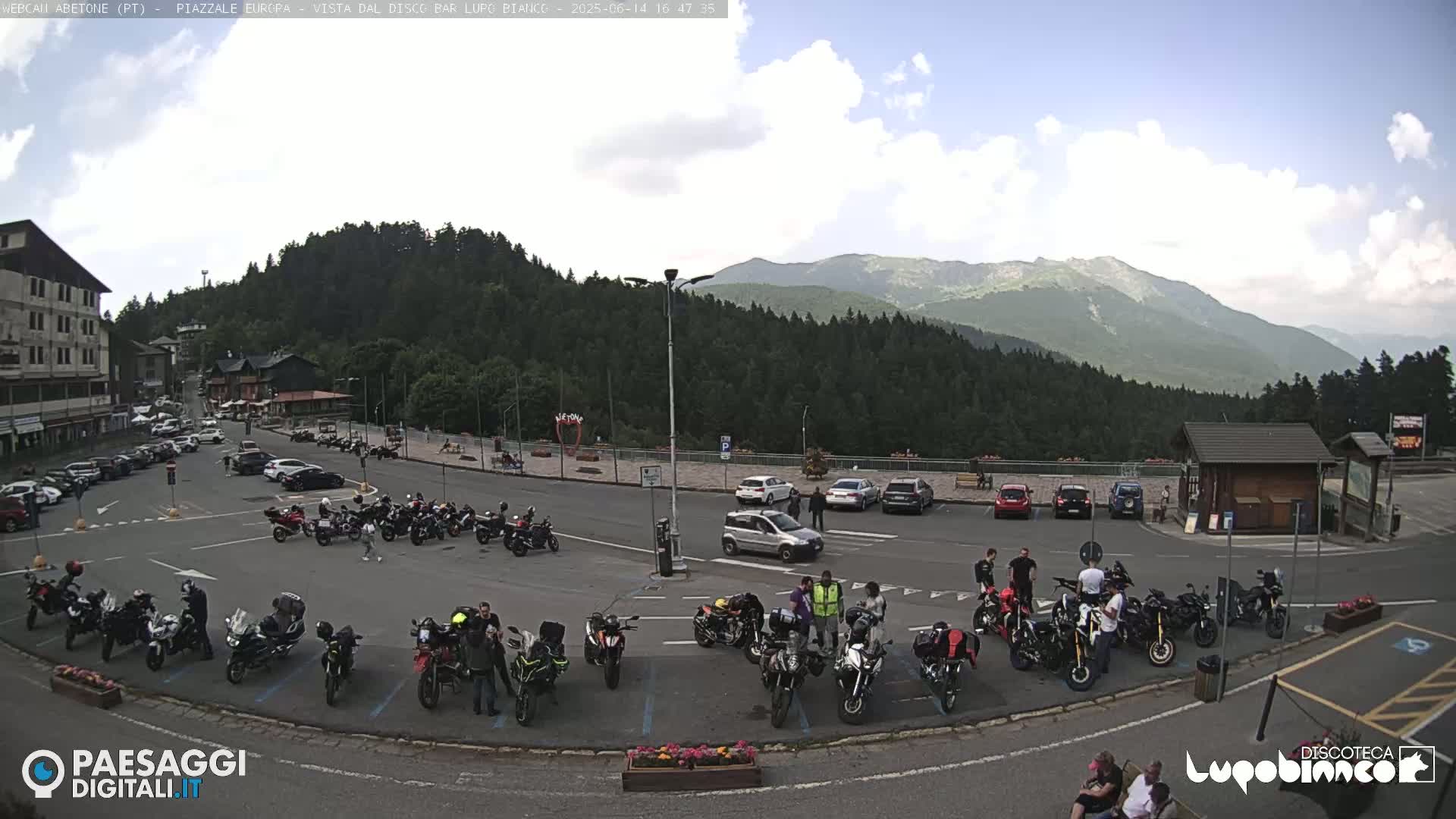 A parking lot filled with motorcycles and cars is situated in front of a mountain range under a partly cloudy sky.