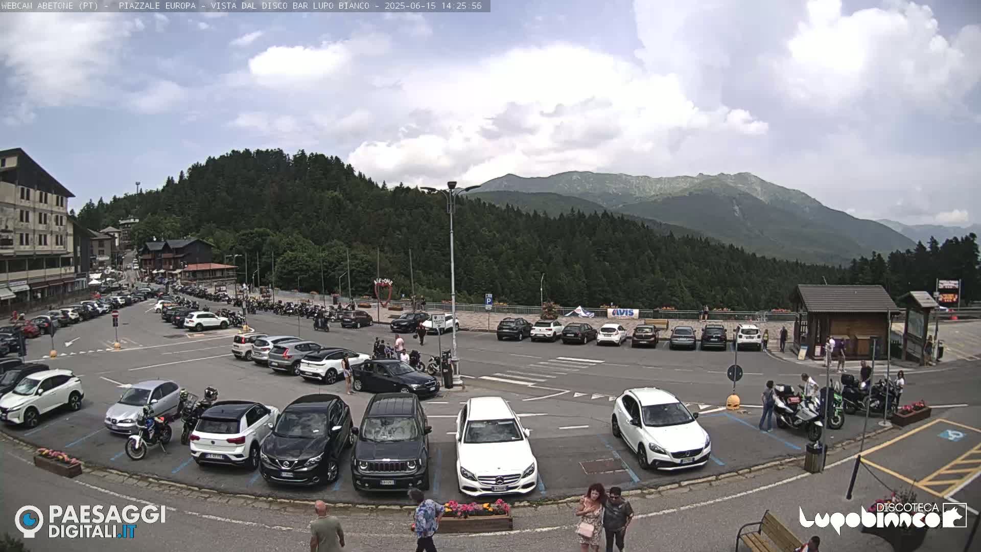 A parking lot filled with cars and motorcycles is situated in front of a mountain range under a partly cloudy sky.