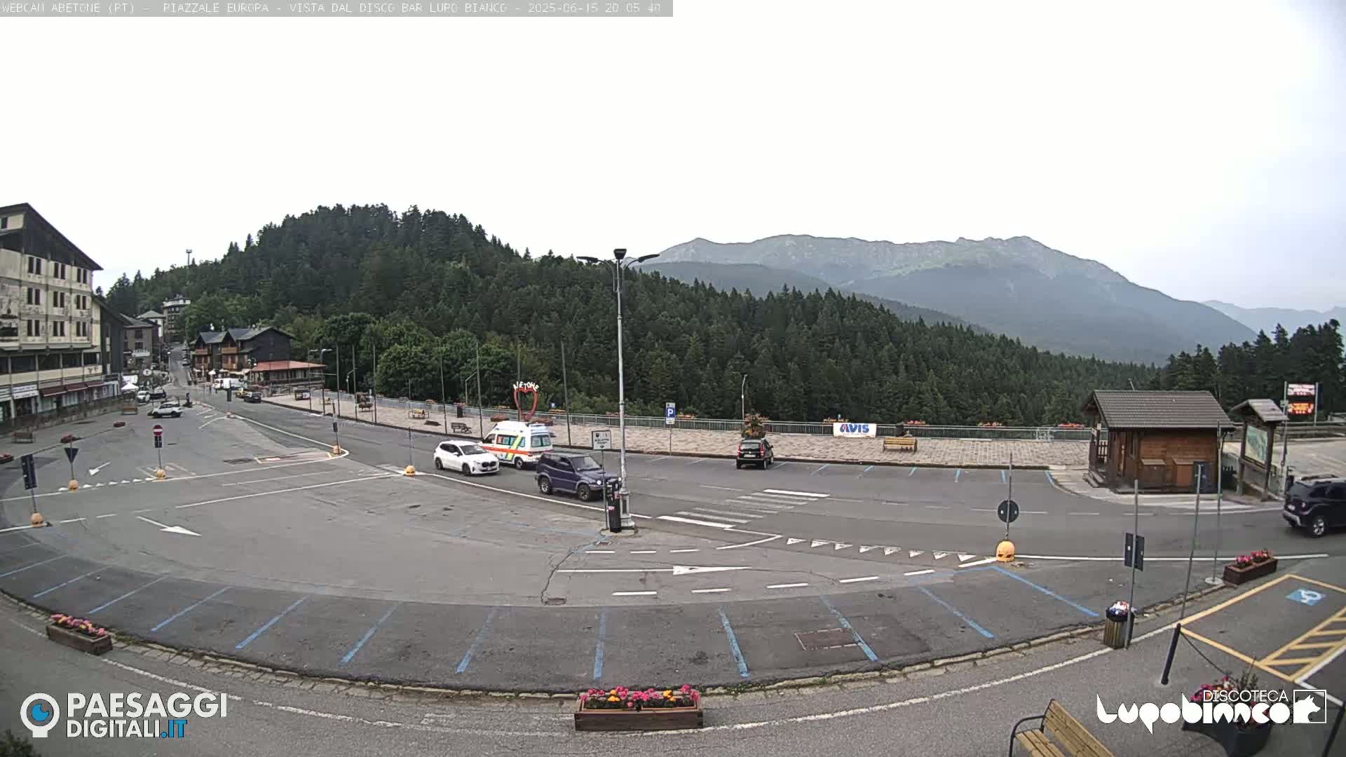 A mostly empty parking lot with several cars and a medical vehicle is situated in a mountain town, with a mountain range visible in the background under an overcast sky.