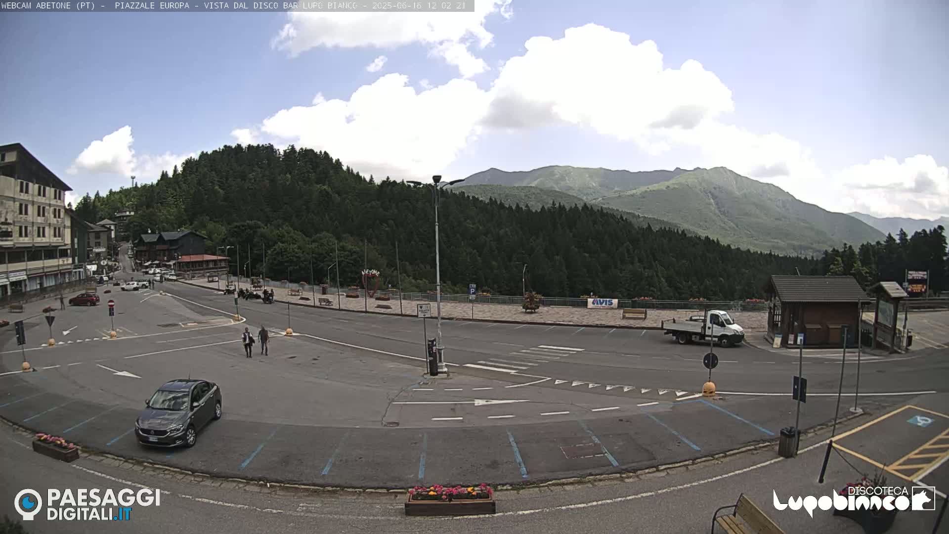 A mostly empty parking lot and roadway in a mountain town under a partly cloudy sky, with a few cars and people visible, and mountains in the background.