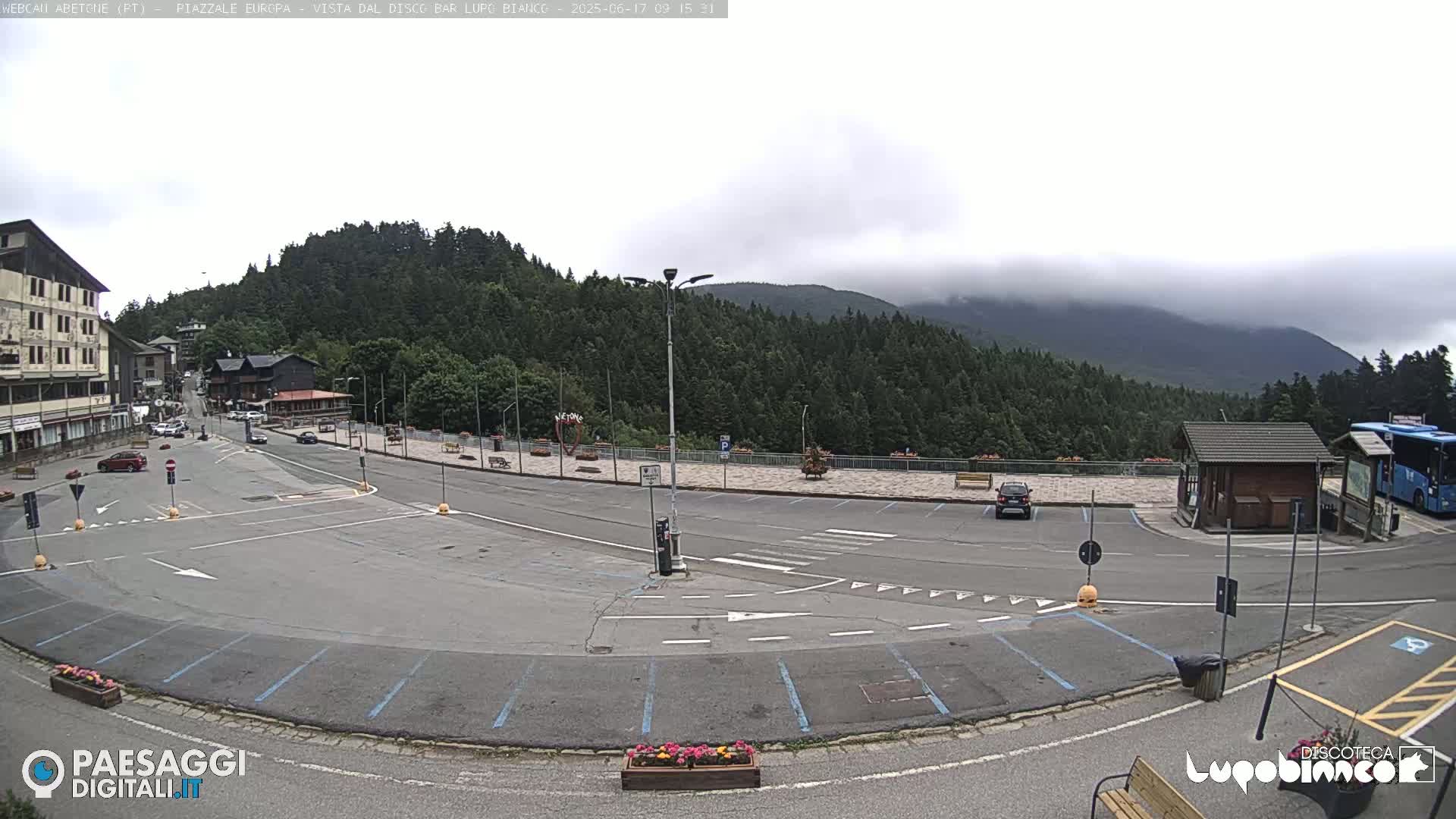 An empty parking lot and roadway are situated in front of buildings, with a forested mountain in the background under an overcast sky.
