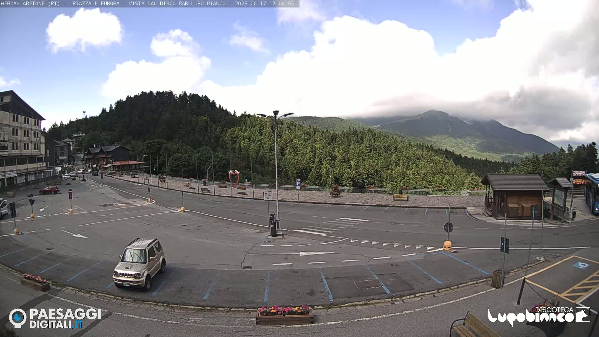 A mostly empty parking lot is situated in a mountainous area under a partly cloudy sky, with a few cars and a small building visible.
