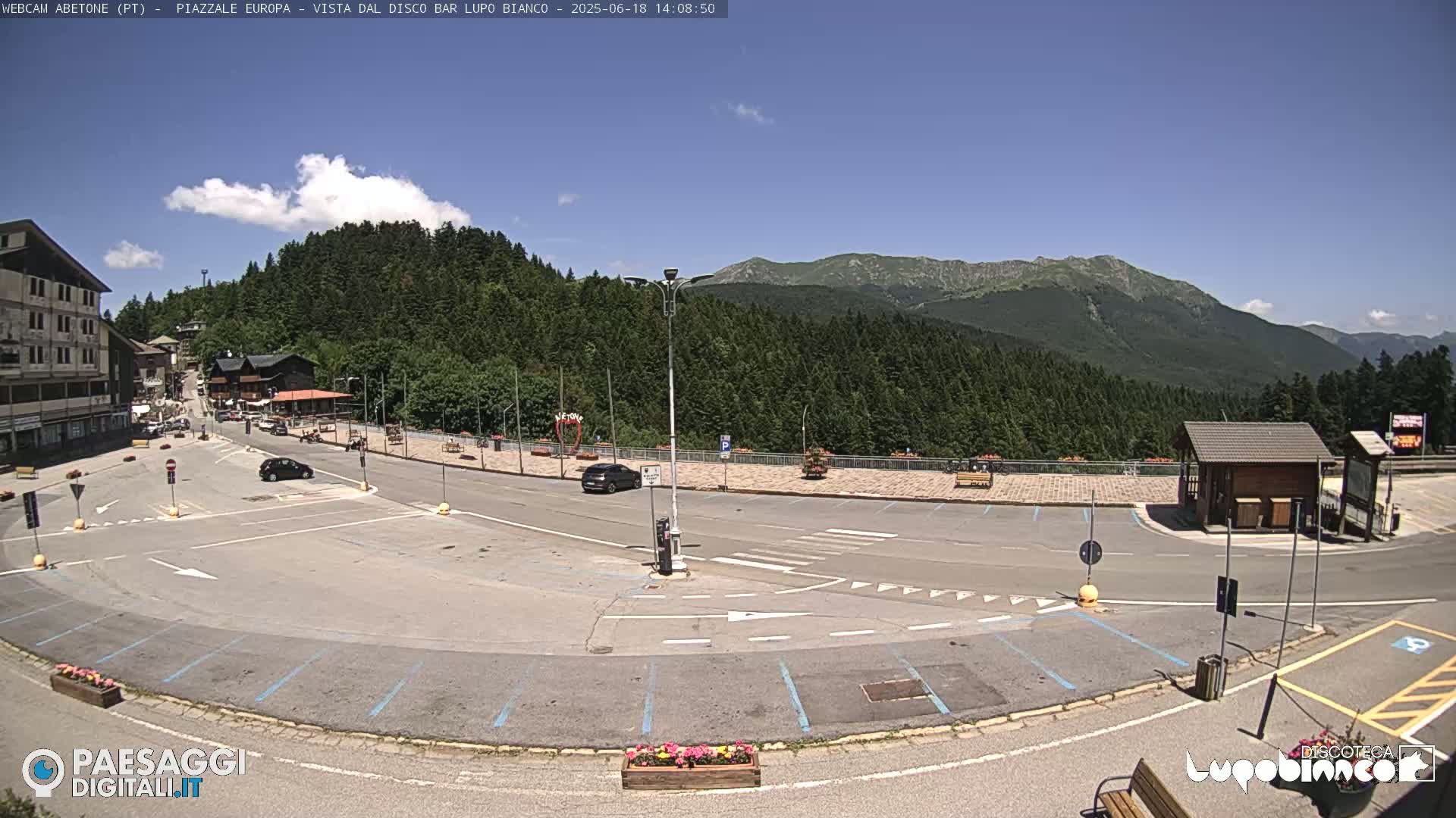 An empty parking lot and roadway in a mountainous area on a sunny day, with a few cars and a small building visible.