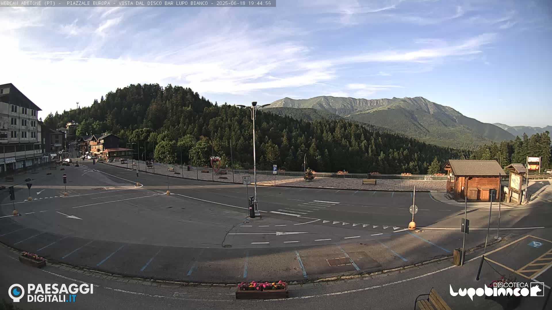 An empty parking lot and roadway are situated in front of a building and forested mountain range under a mostly clear sky.