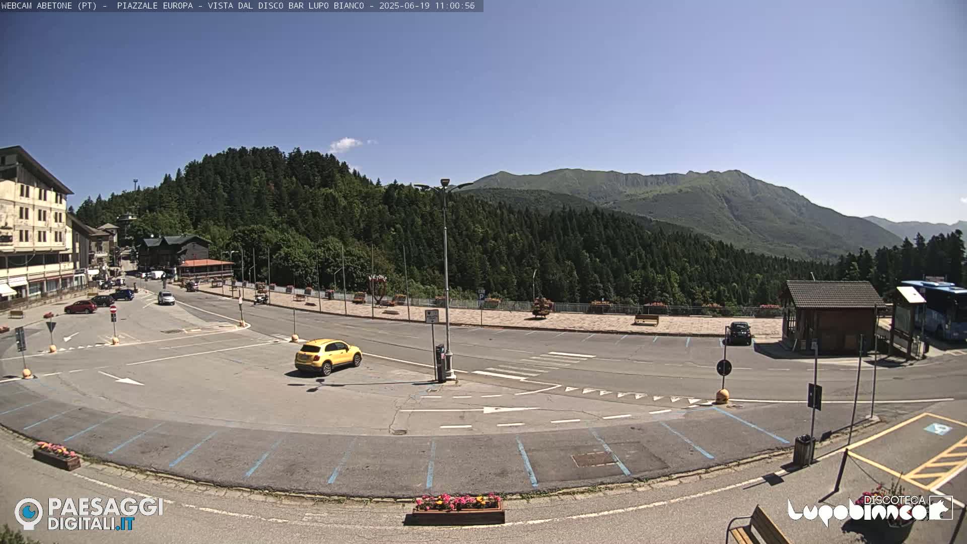 A mostly empty parking lot with a yellow car sits in front of a building in a mountainous area under a clear blue sky.