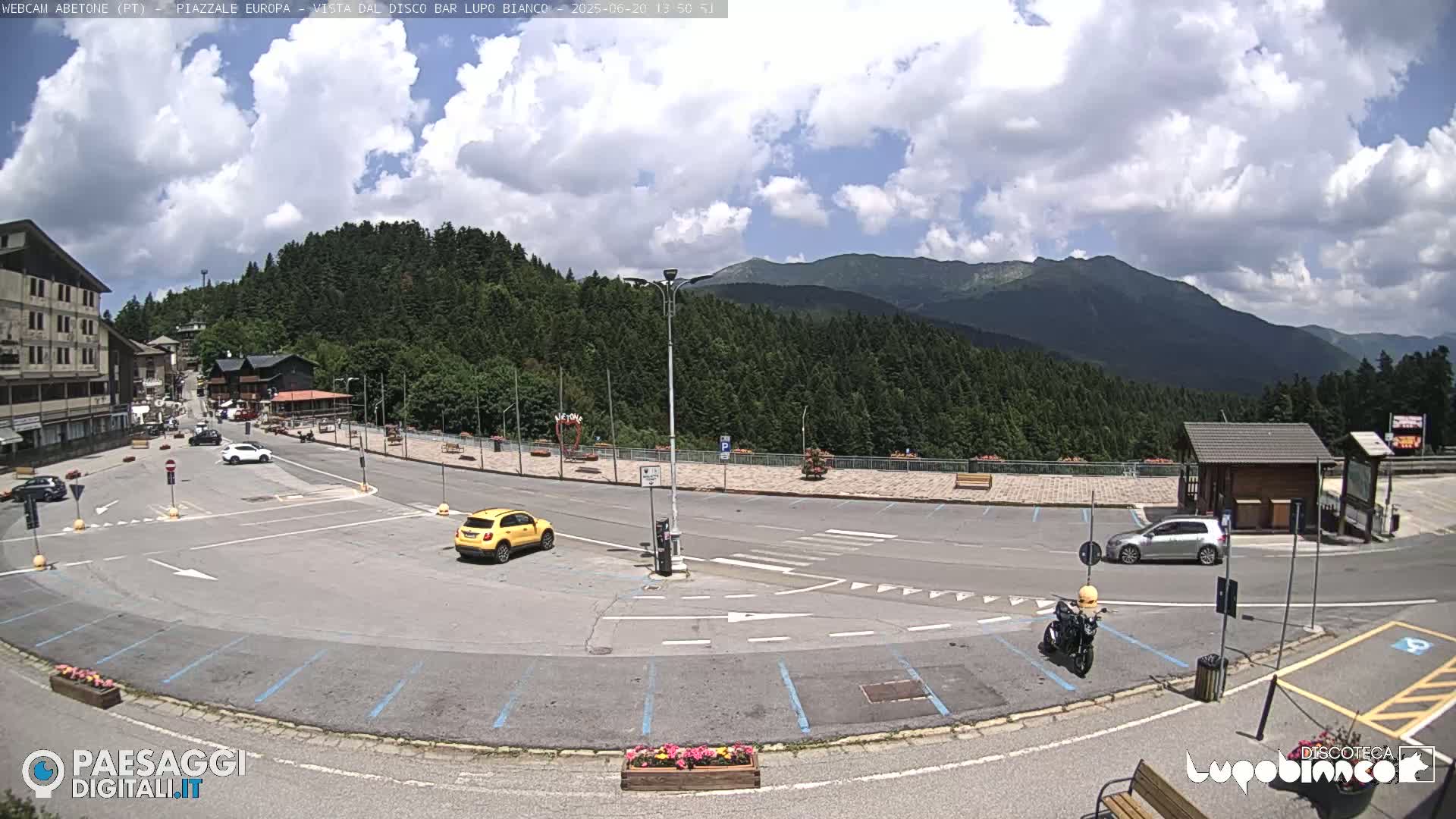 A mostly empty parking lot with several cars and a motorcycle, situated in front of a building and a wooded mountain range under a partly cloudy sky.