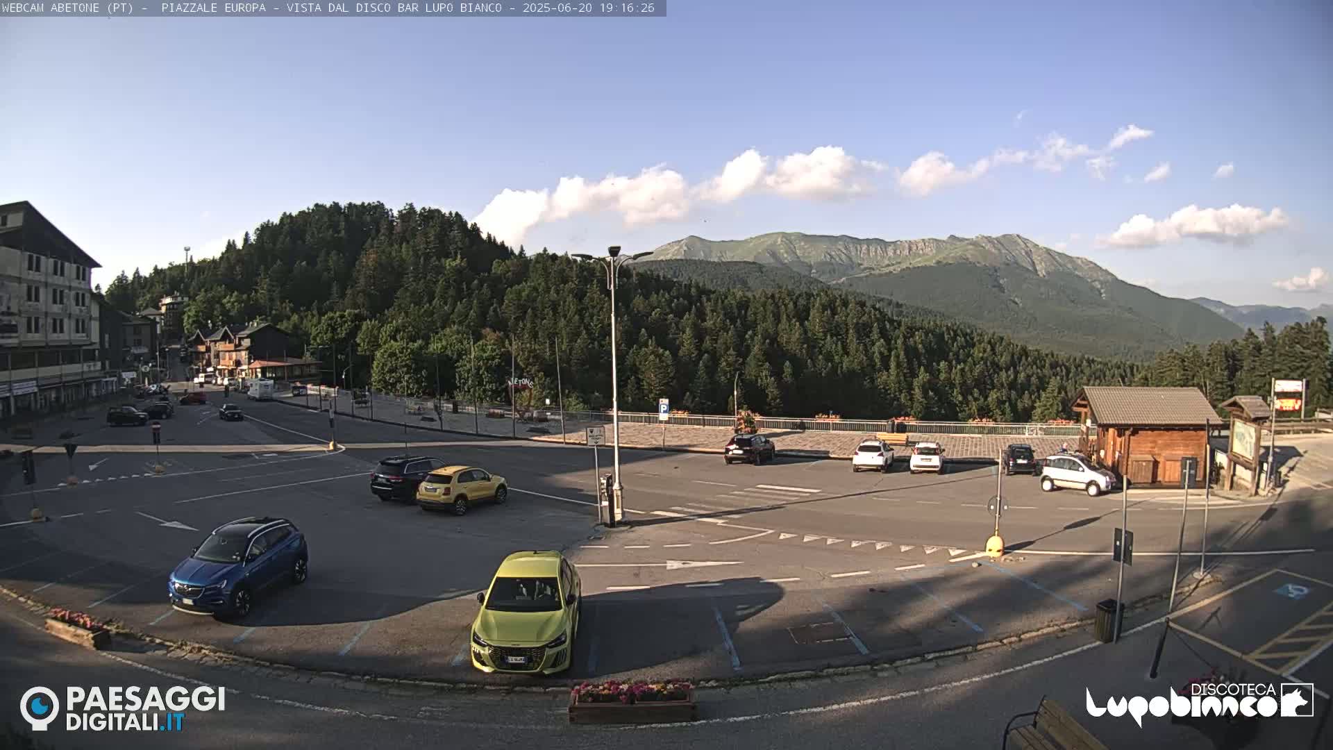 A mostly sunny mountain vista overlooks a parking lot with several cars, a small building, and a road intersection.