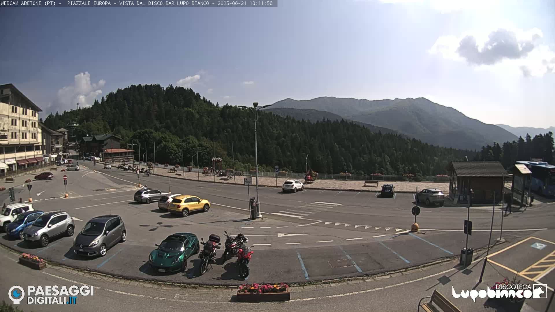 A sunny day reveals a mountain vista overlooking a parking lot with several cars and motorcycles parked near a building, with a few people visible in the distance.