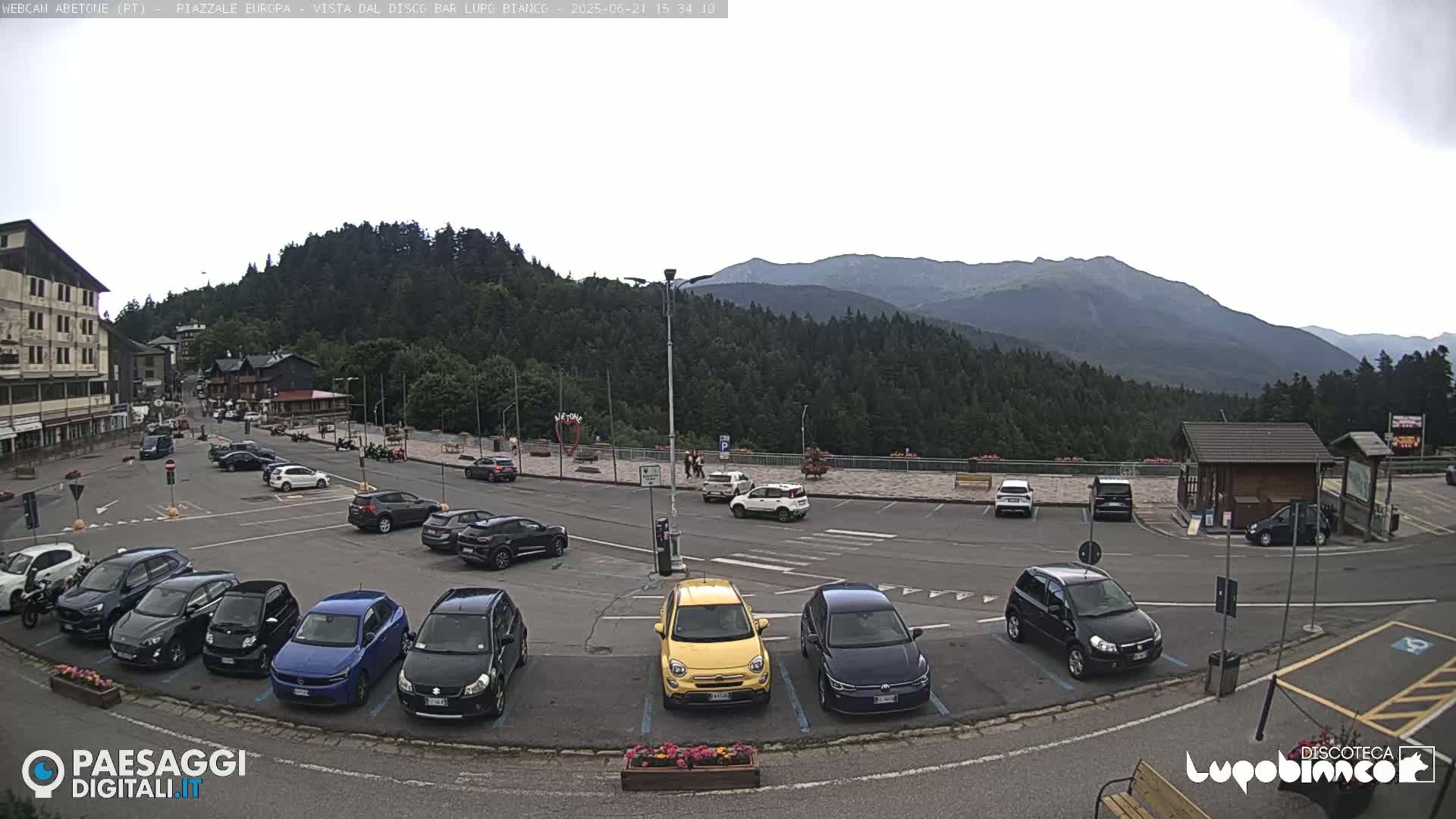 A parking lot filled with cars is situated in front of a building and overlooks a mountain range under an overcast sky.