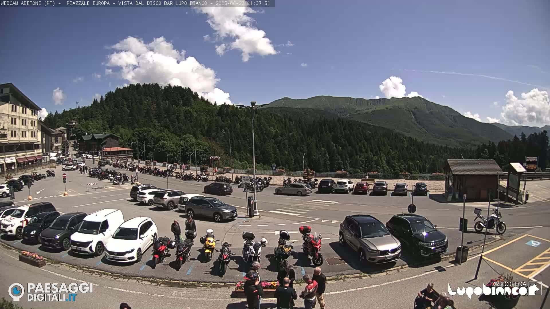 A sunny day in a mountain parking lot filled with cars and motorcycles, with a backdrop of lush green hills and a clear blue sky.