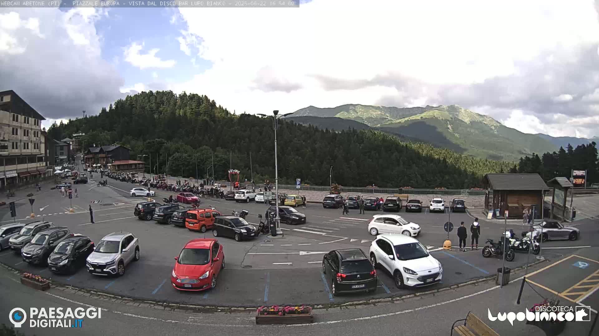 A parking lot filled with cars and motorcycles is situated in a mountainous area under partly cloudy skies.