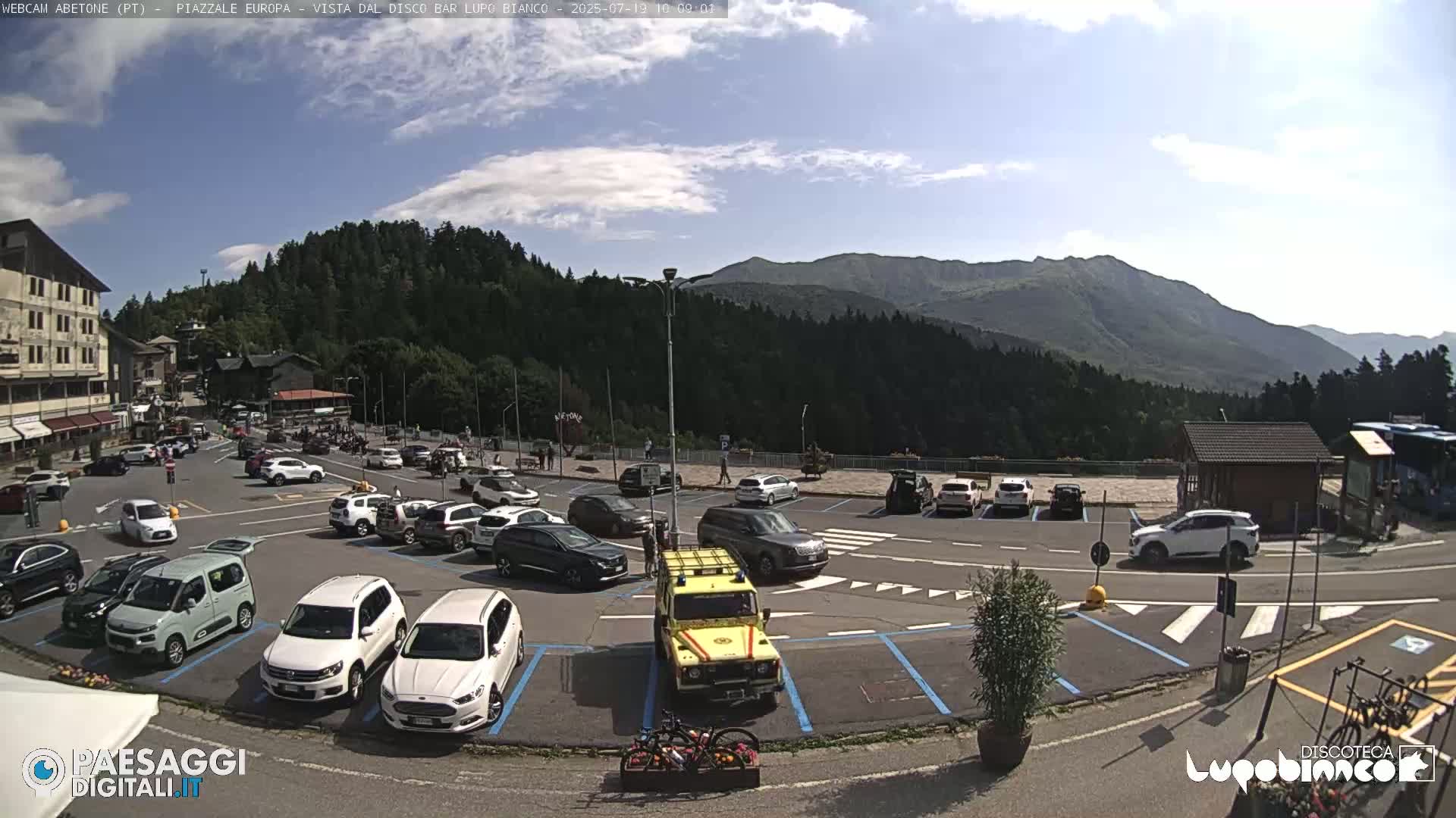 A sunny day reveals a mountain view overlooking a parking lot filled with cars, a small building, and some bicycles.
