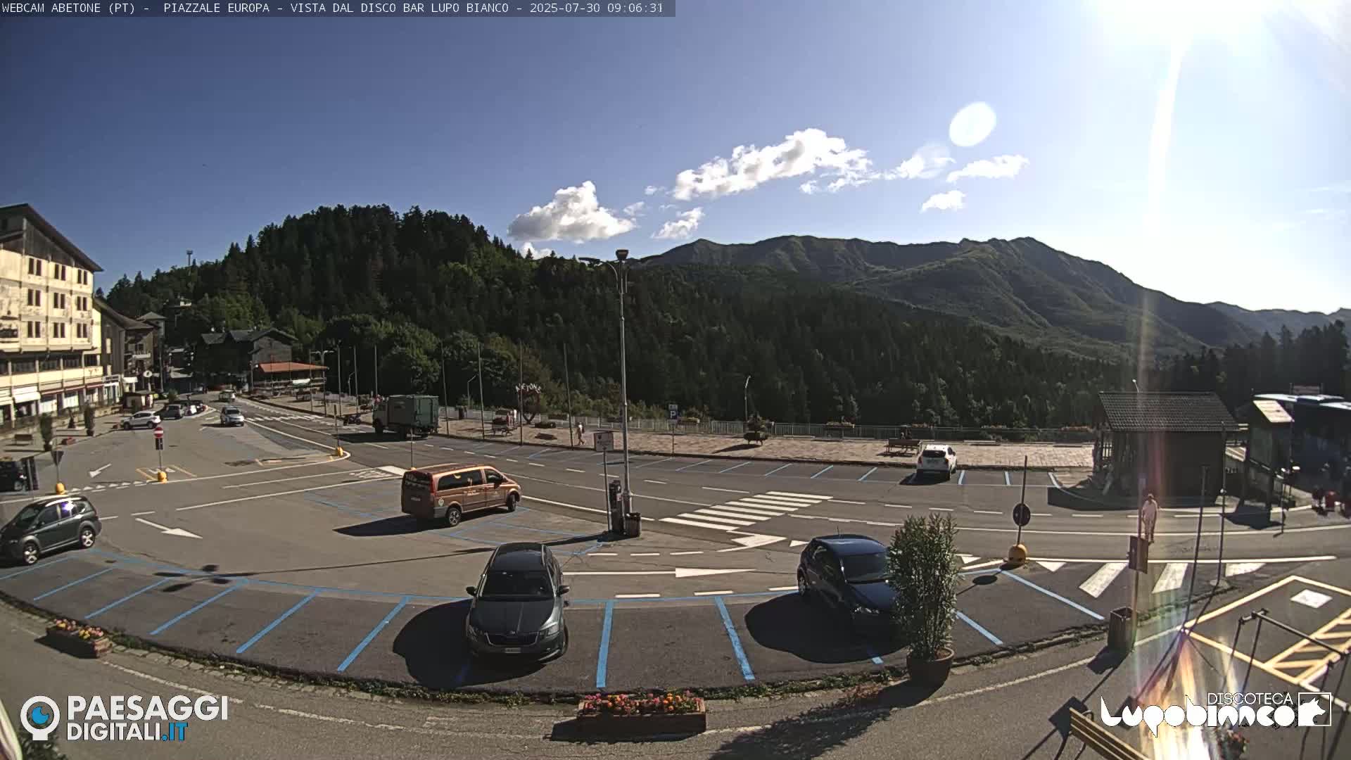 A mountainside parking lot with several cars, a van, and a building, under a mostly sunny sky with a few clouds.