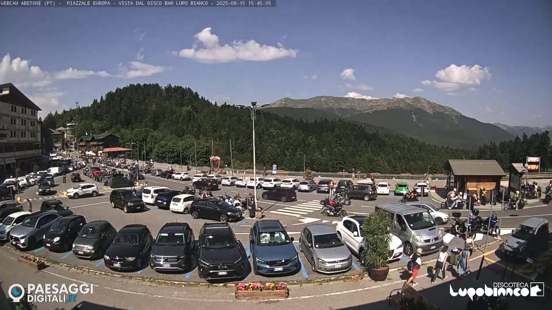 A mountainside parking lot with several cars, a van, and a building, under a mostly sunny sky with a few clouds.