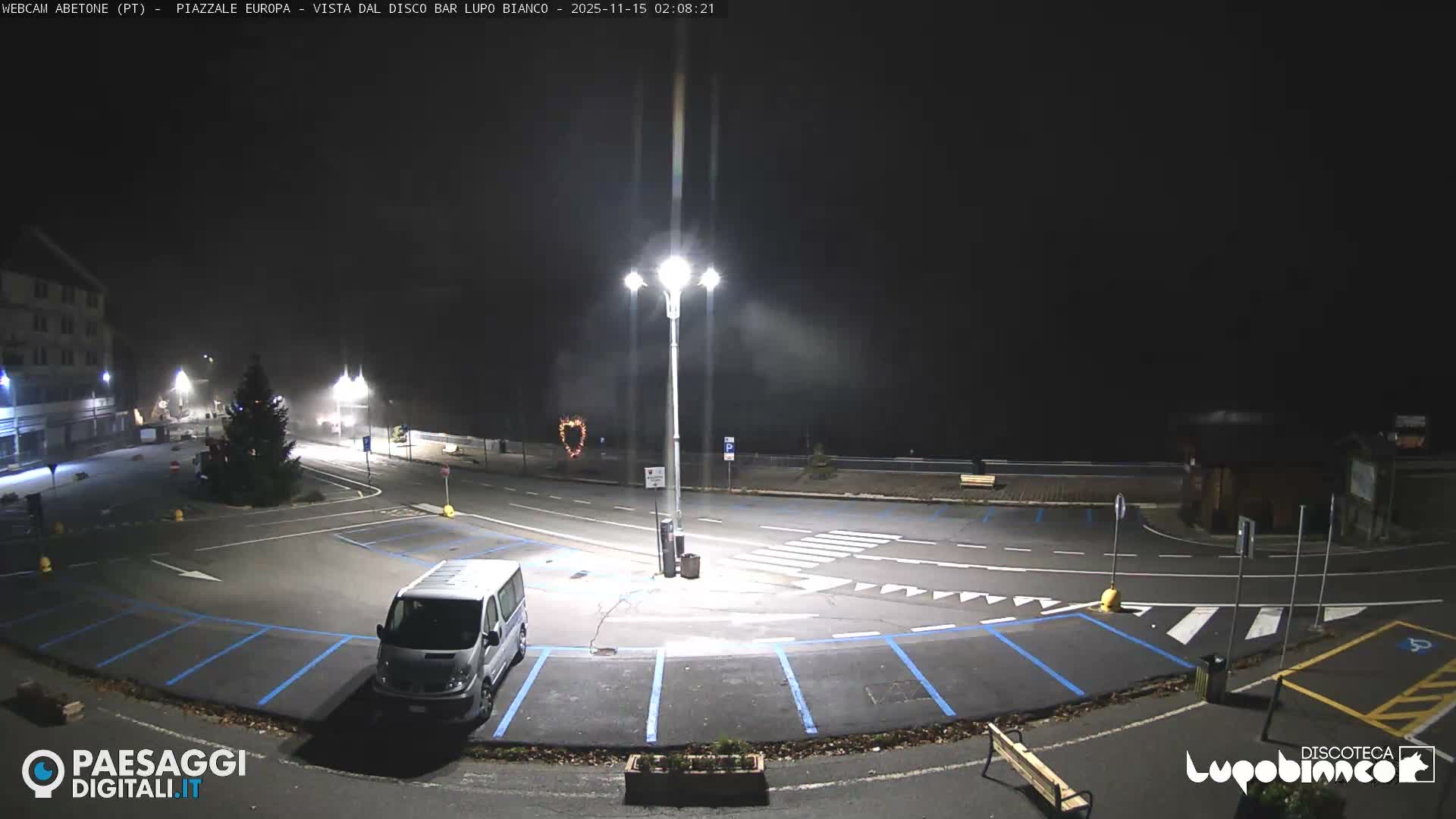 A foggy night view captures an illuminated town square featuring a decorated festive tree, a prominent streetlight, a parked silver van, and a heart-shaped light decoration.