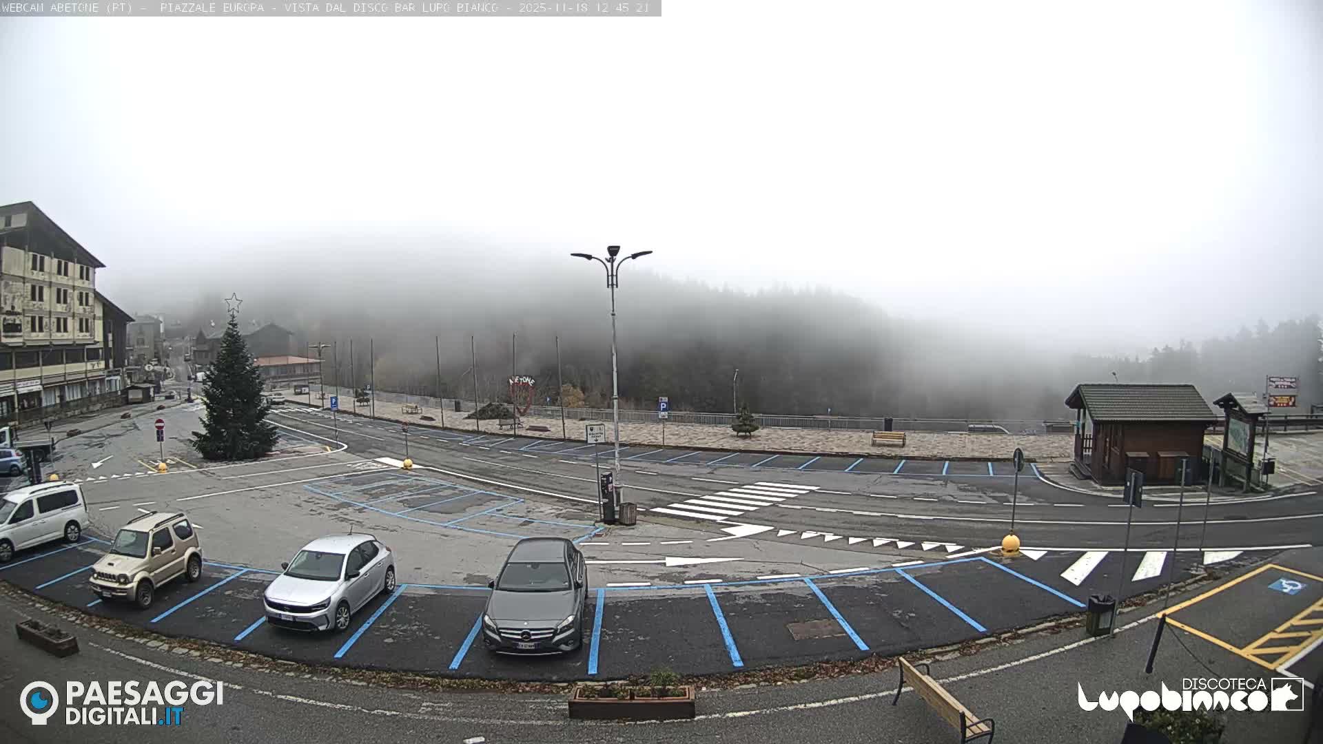 A foggy night view captures an illuminated town square featuring a decorated festive tree, a prominent streetlight, a parked silver van, and a heart-shaped light decoration.