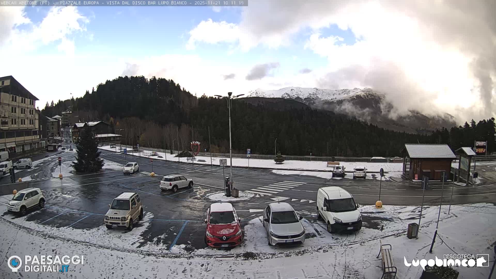 A snow-covered alpine town scene unfolds under a partly cloudy sky, featuring a central road and parking area with several cars, surrounded by multi-story buildings, dense pine forests, and distant snow-capped mountains partially veiled in mist.