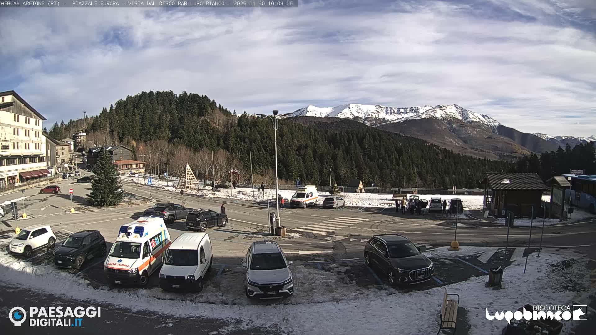 A partly cloudy winter day reveals a bustling mountain town square with numerous cars and an ambulance parked on partially snow-dusted ground, surrounded by buildings, a dense pine forest, and distant snow-capped mountains.