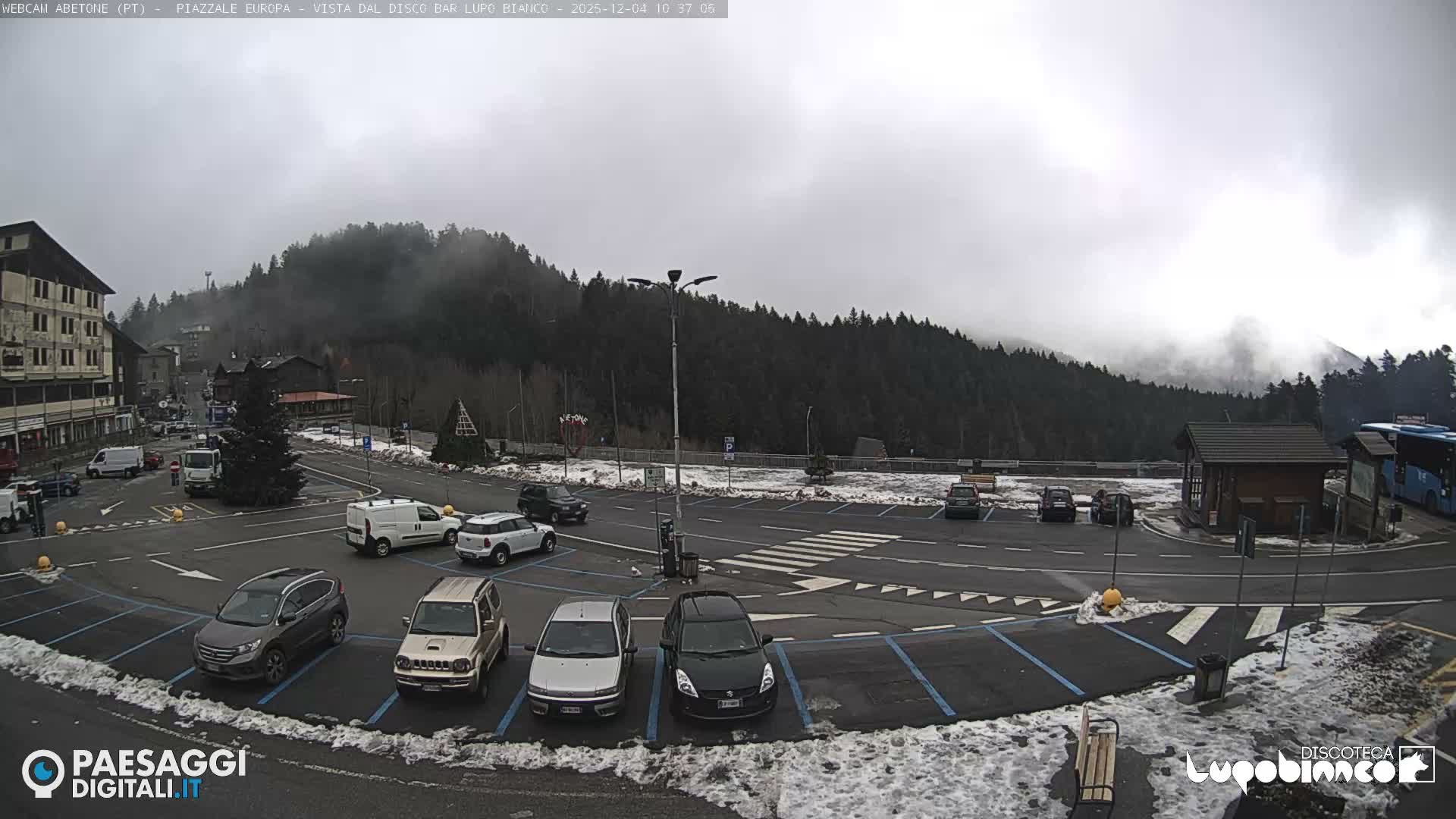 The image depicts a cloudy winter day in a mountain town, showing a street, a snow-dusted parking lot with various vehicles, buildings, a decorated Christmas tree, and forested hills partially shrouded in mist.
