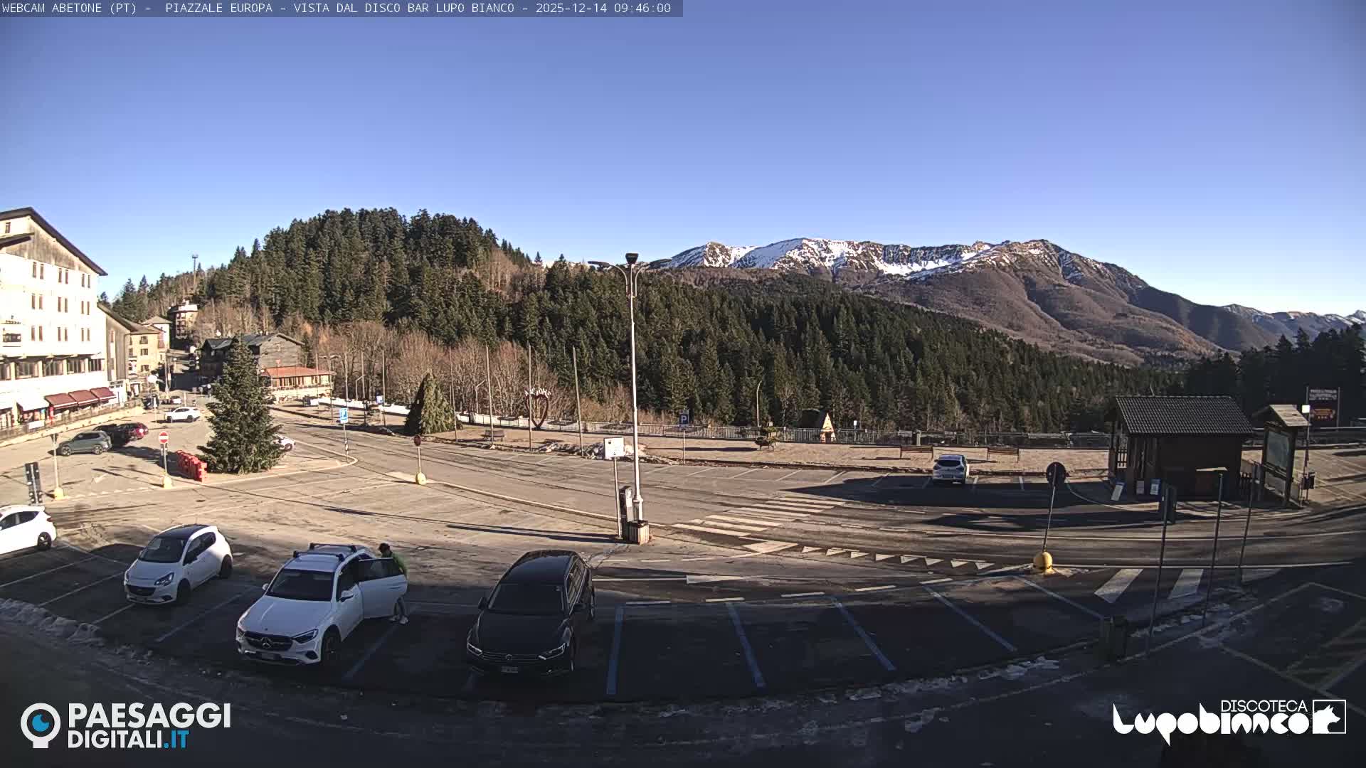 The image depicts a cloudy winter day in a mountain town, showing a street, a snow-dusted parking lot with various vehicles, buildings, a decorated Christmas tree, and forested hills partially shrouded in mist.
