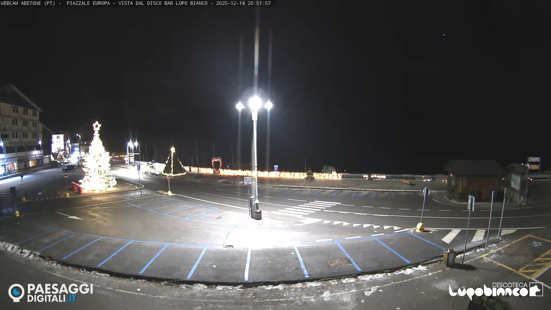 The image depicts a cloudy winter day in a mountain town, showing a street, a snow-dusted parking lot with various vehicles, buildings, a decorated Christmas tree, and forested hills partially shrouded in mist.