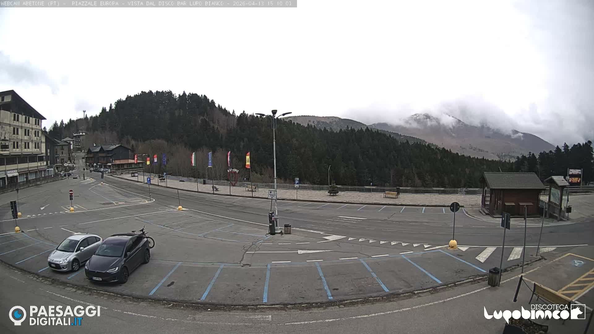 The image depicts a cloudy winter day in a mountain town, showing a street, a snow-dusted parking lot with various vehicles, buildings, a decorated Christmas tree, and forested hills partially shrouded in mist.