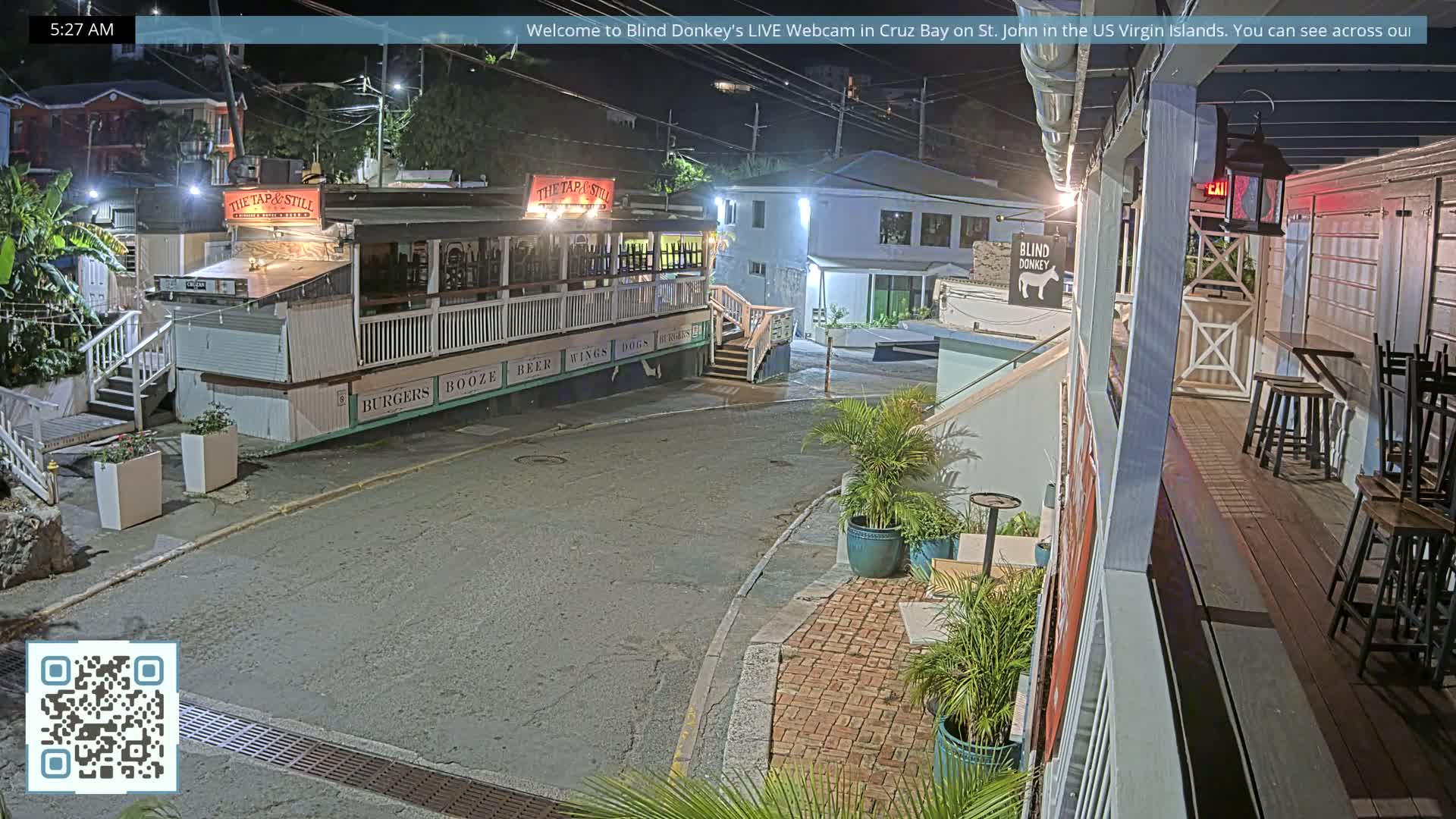 The image depicts a quiet, artificially lit street scene at early morning with a multi-story building featuring outdoor seating on the left and other structures on the right, all under a clear dark sky.