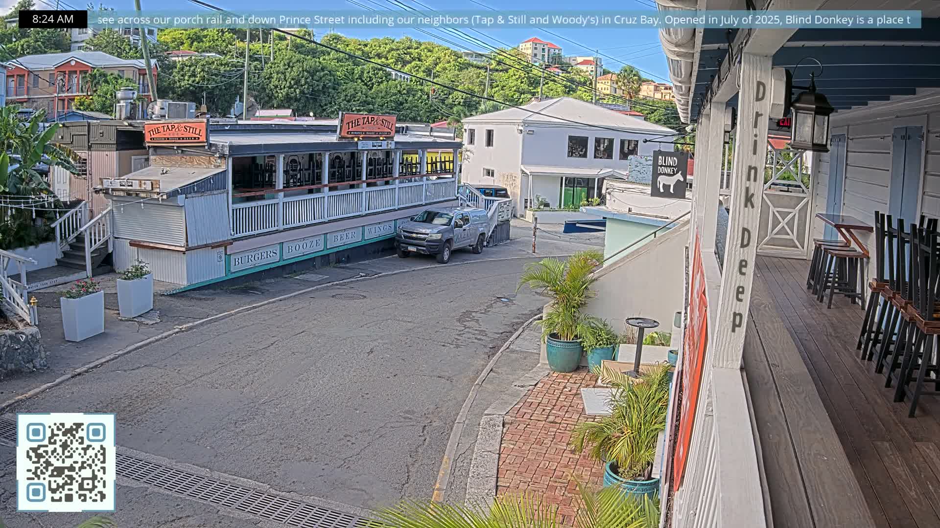 The image depicts a quiet, artificially lit street scene at early morning with a multi-story building featuring outdoor seating on the left and other structures on the right, all under a clear dark sky.