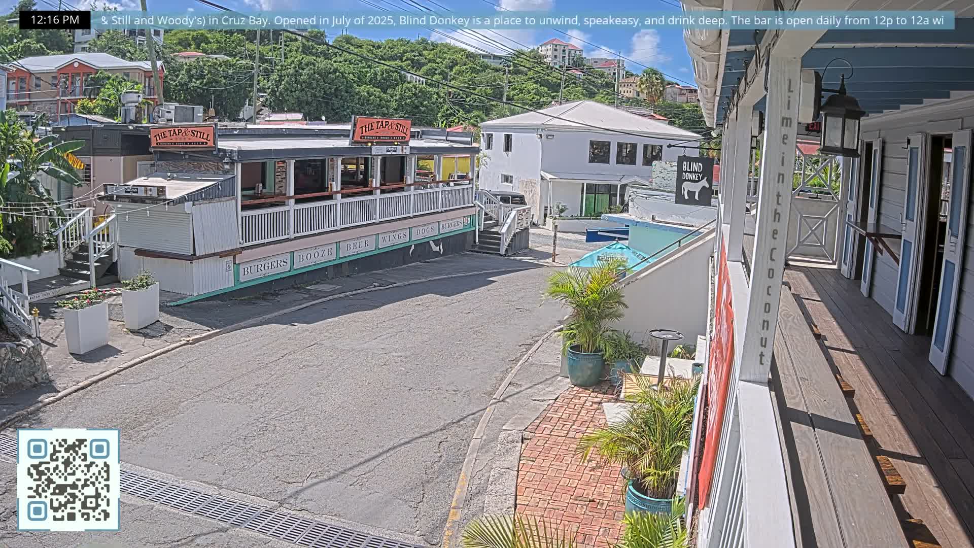 Under a bright, sunny sky, a quiet street with brick and concrete sidewalks winds between buildings, one featuring a large open-air balcony, and ascends towards lush, green hills in the background.