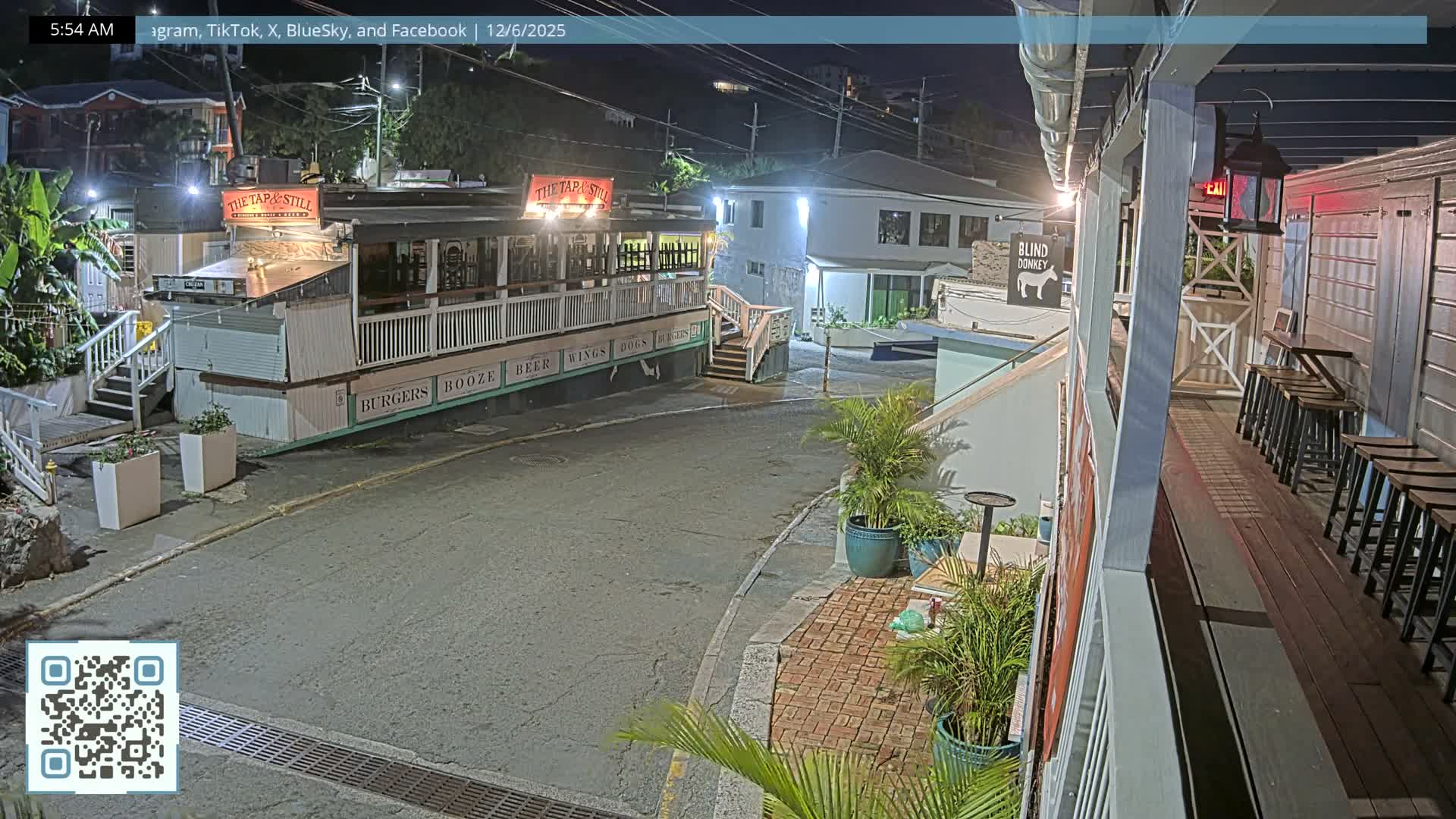 An outdoor nocturnal scene depicts a quiet, artificially lit street in a town, featuring a prominent bar-like building with a large outdoor deck and multiple sets of steps, alongside other structures, adorned with numerous potted plants, all under clear skies.