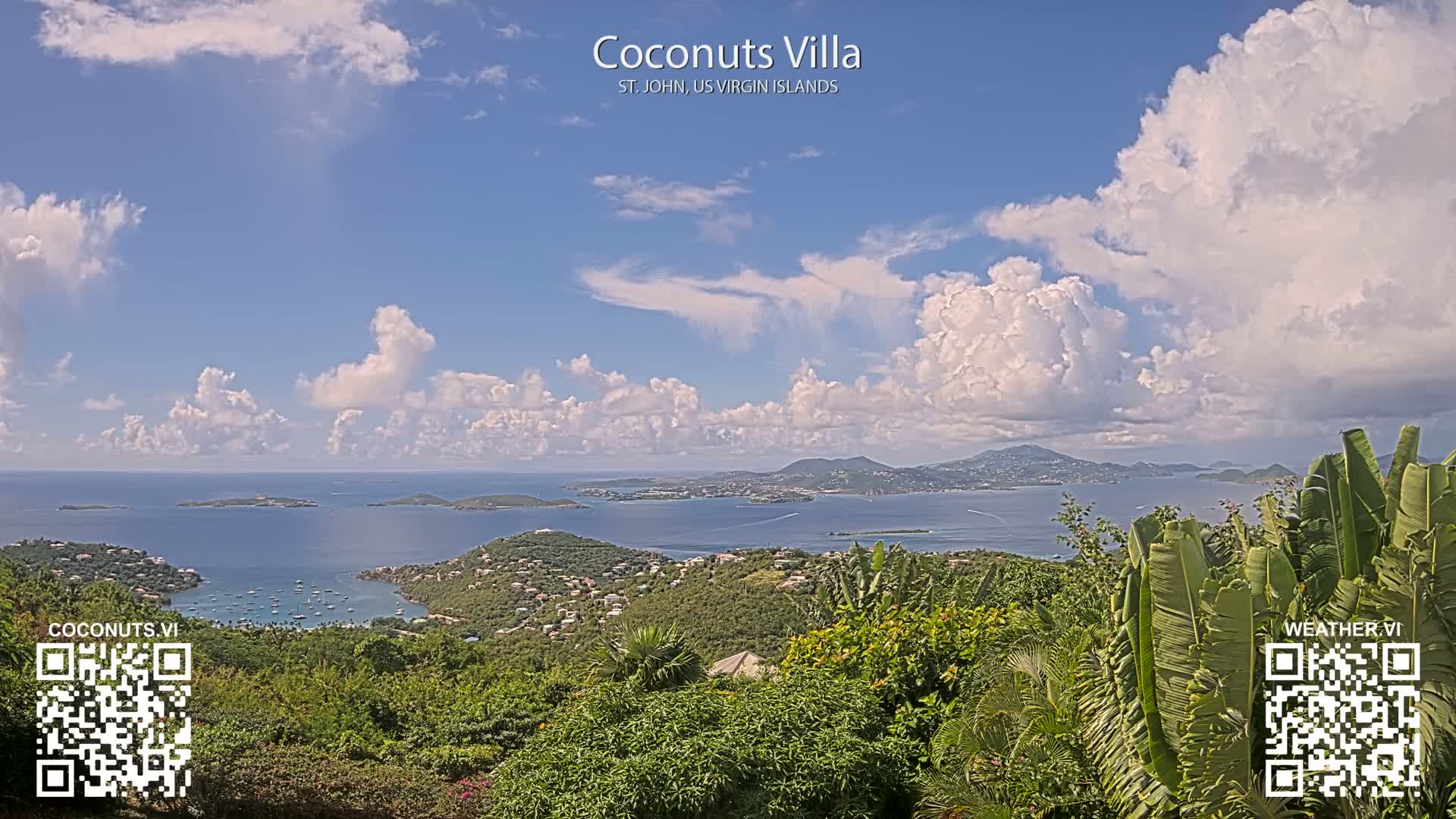 Under a fair weather sky with scattered white clouds, a panoramic view reveals lush green hills dotted with buildings, a tranquil bay filled with anchored boats, and numerous islands stretching across the blue ocean to distant mountains.