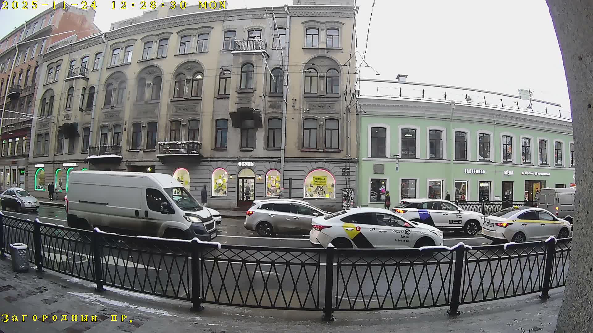 On an overcast and snowy day, an urban street features historic-looking buildings with ground-floor shops, several cars and taxis driving on a wet road, and a few pedestrians on the sidewalks.