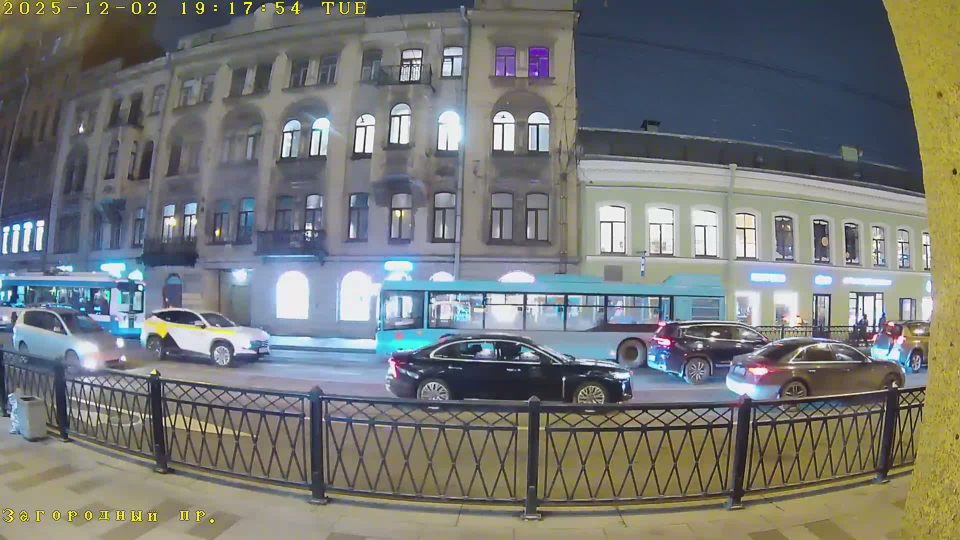 A clear night scene shows an urban street bustling with cars and buses in traffic, illuminated by streetlights and the bright windows of multi-story buildings lining the road.