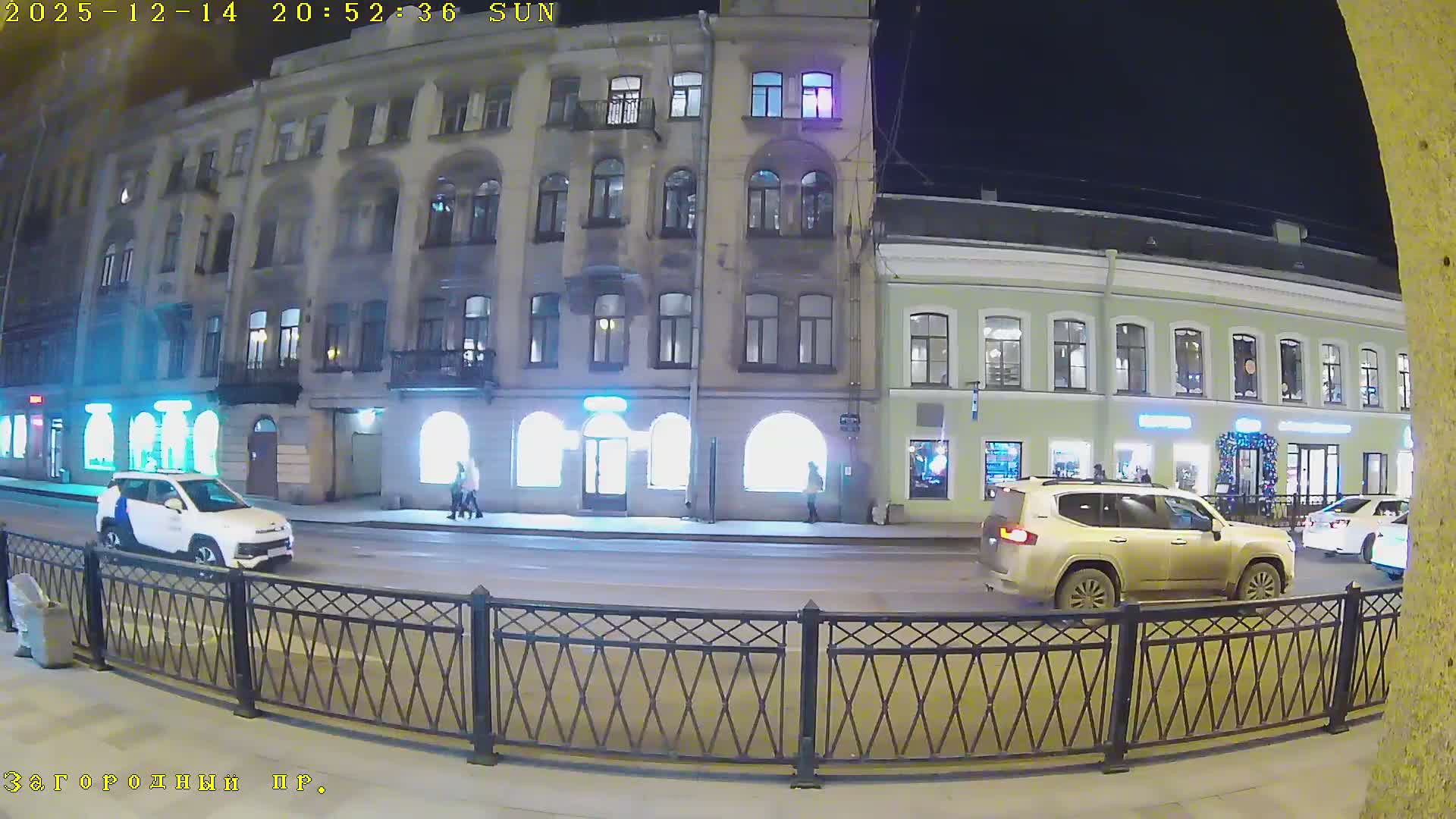 An overcast urban street scene shows vehicular traffic moving along a multi-lane road bordered by historic multi-story buildings with ground-floor shops and a few pedestrians on the sidewalks.