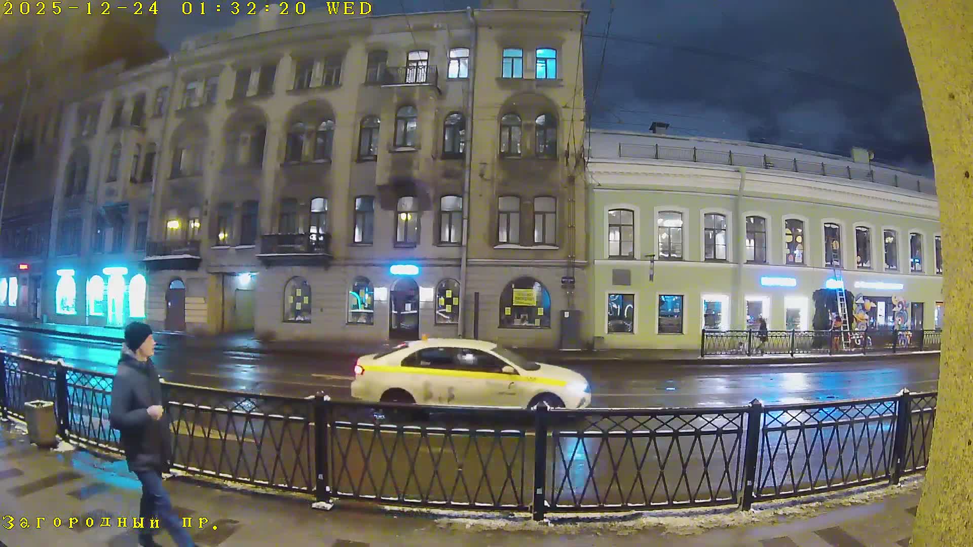 An overcast urban street scene shows vehicular traffic moving along a multi-lane road bordered by historic multi-story buildings with ground-floor shops and a few pedestrians on the sidewalks.