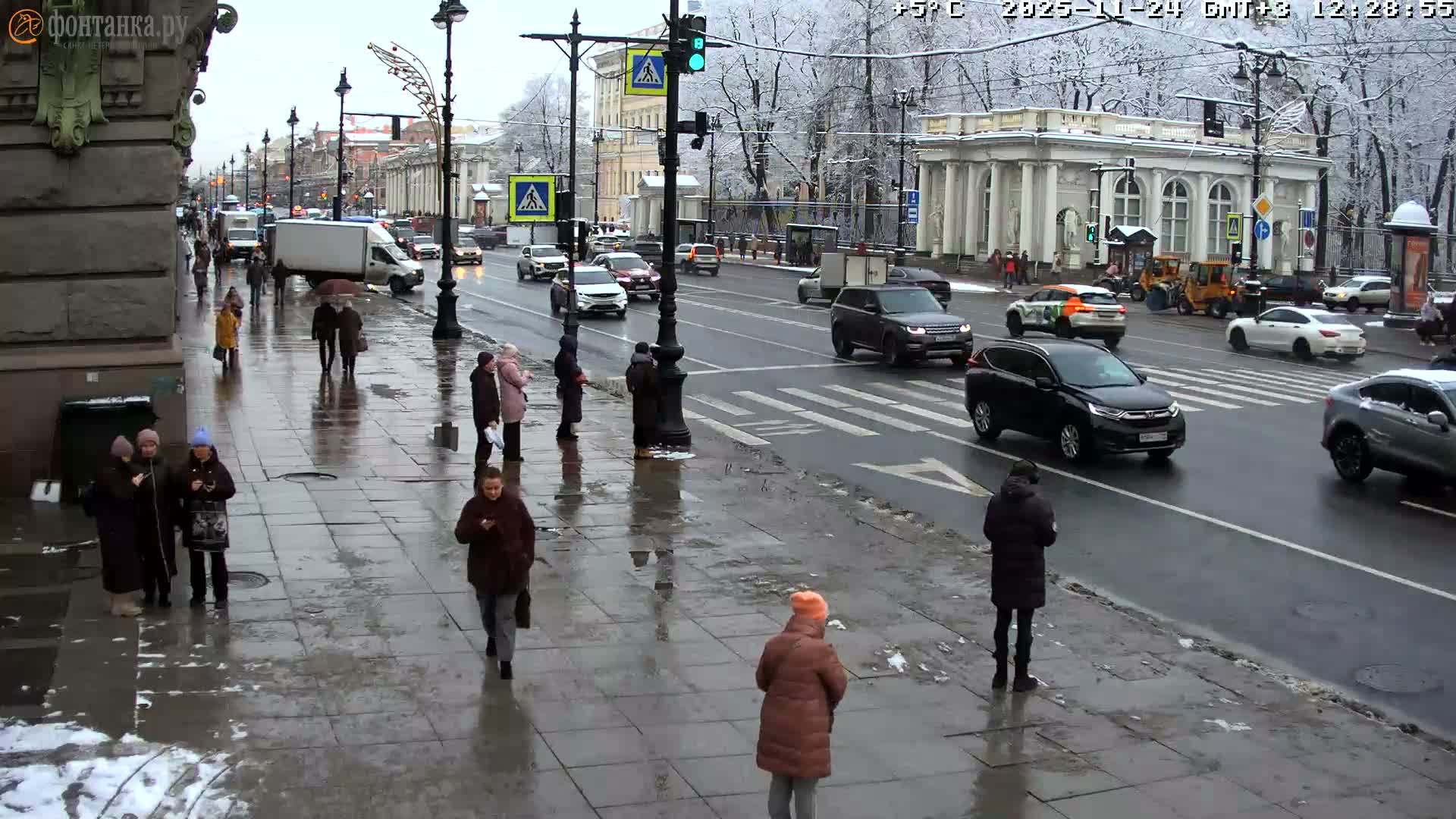 Pedestrians navigate wet sidewalks alongside a busy street with cars and trucks, under an overcast winter sky with melting snow clinging to bare trees.