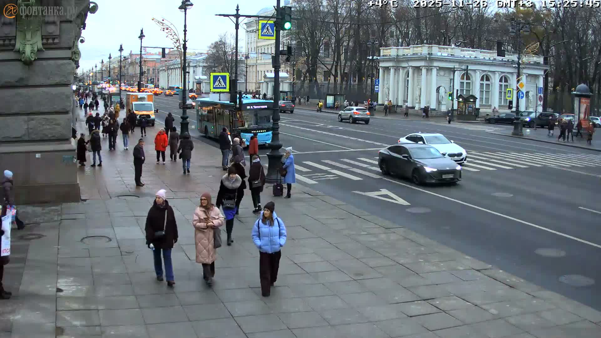 Pedestrians navigate wet sidewalks alongside a busy street with cars and trucks, under an overcast winter sky with melting snow clinging to bare trees.
