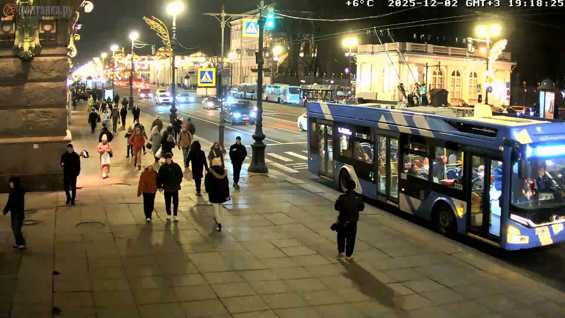 A bustling city street at night shows numerous pedestrians walking on a tiled sidewalk, a large blue and white trolleybus in the foreground, and other vehicles on the road, all illuminated by streetlights under a clear +6°C sky.