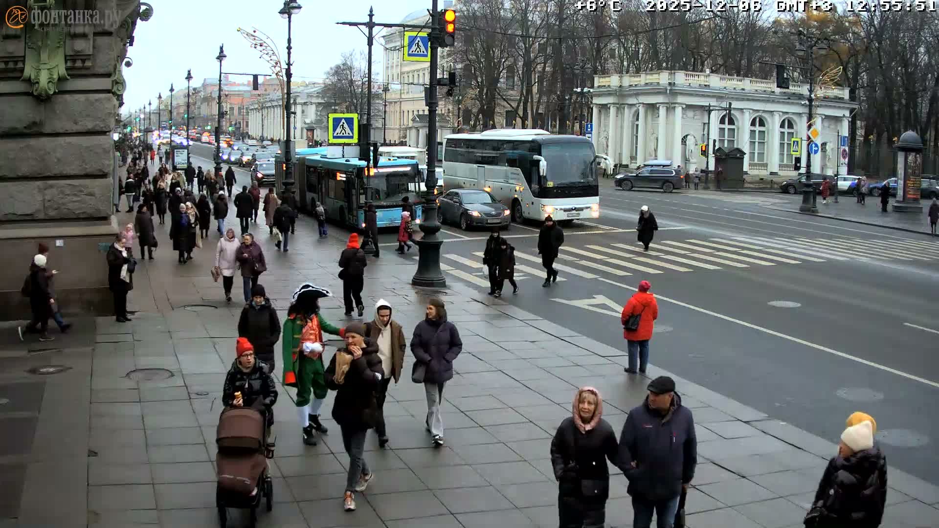 A bustling urban street is depicted on a chilly, overcast, and wet day, with numerous pedestrians, including one in historical costume, traversing sidewalks and crosswalks alongside cars and buses on the damp road, framed by classic buildings and bare trees.