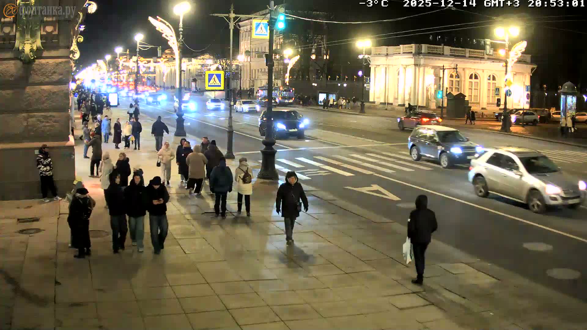 A bustling urban street is depicted on a chilly, overcast, and wet day, with numerous pedestrians, including one in historical costume, traversing sidewalks and crosswalks alongside cars and buses on the damp road, framed by classic buildings and bare trees.