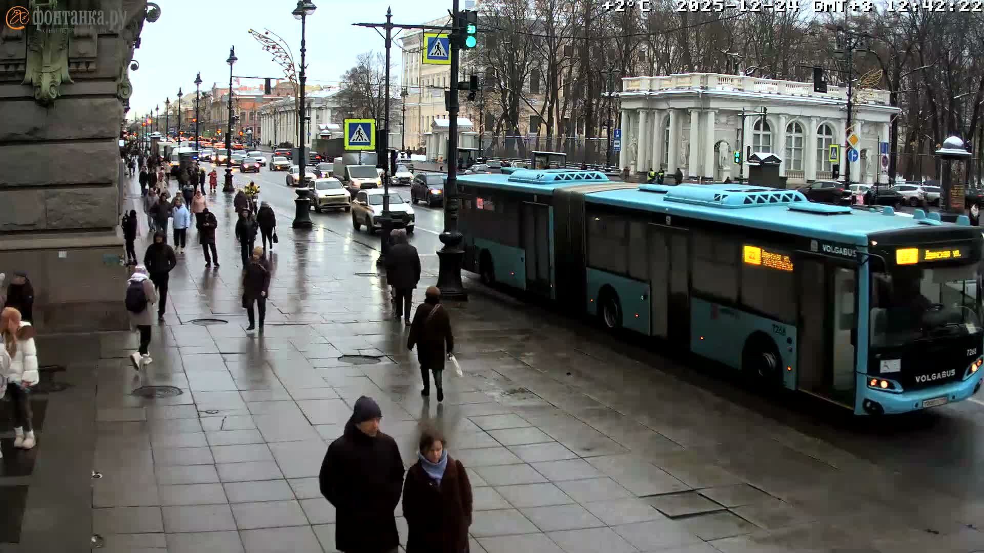 A bustling urban street is depicted on a chilly, overcast, and wet day, with numerous pedestrians, including one in historical costume, traversing sidewalks and crosswalks alongside cars and buses on the damp road, framed by classic buildings and bare trees.