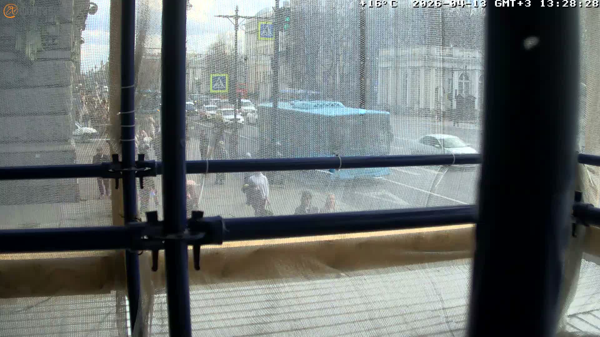 A bustling urban street is depicted on a chilly, overcast, and wet day, with numerous pedestrians, including one in historical costume, traversing sidewalks and crosswalks alongside cars and buses on the damp road, framed by classic buildings and bare trees.