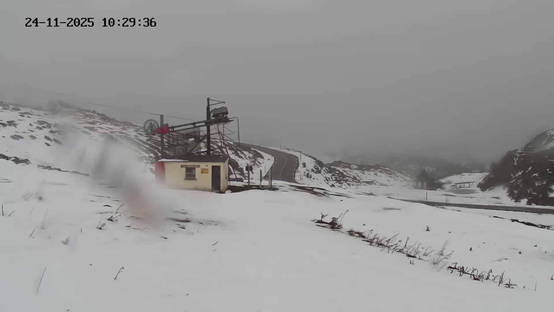A snow-covered mountain landscape is visible under an overcast and misty sky, featuring a small building with a ski lift mechanism above it, a winding road through the snow, and a visible plume of snow being blown across the left foreground.