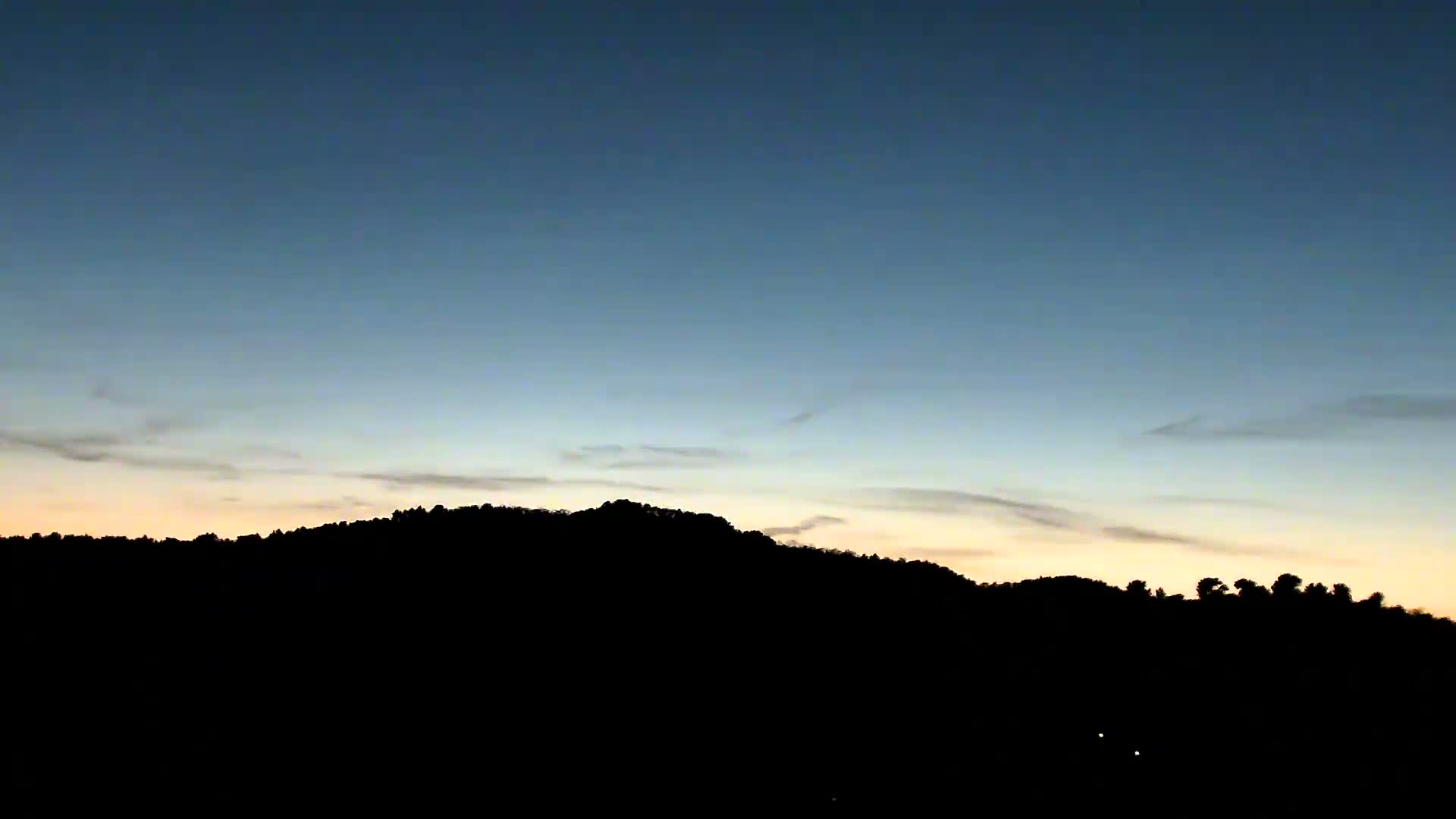 A silhouette of tree-covered mountains is set against a partly cloudy twilight sky, displaying a gradient from deep blue at the top to pale yellow and orange near the horizon.
