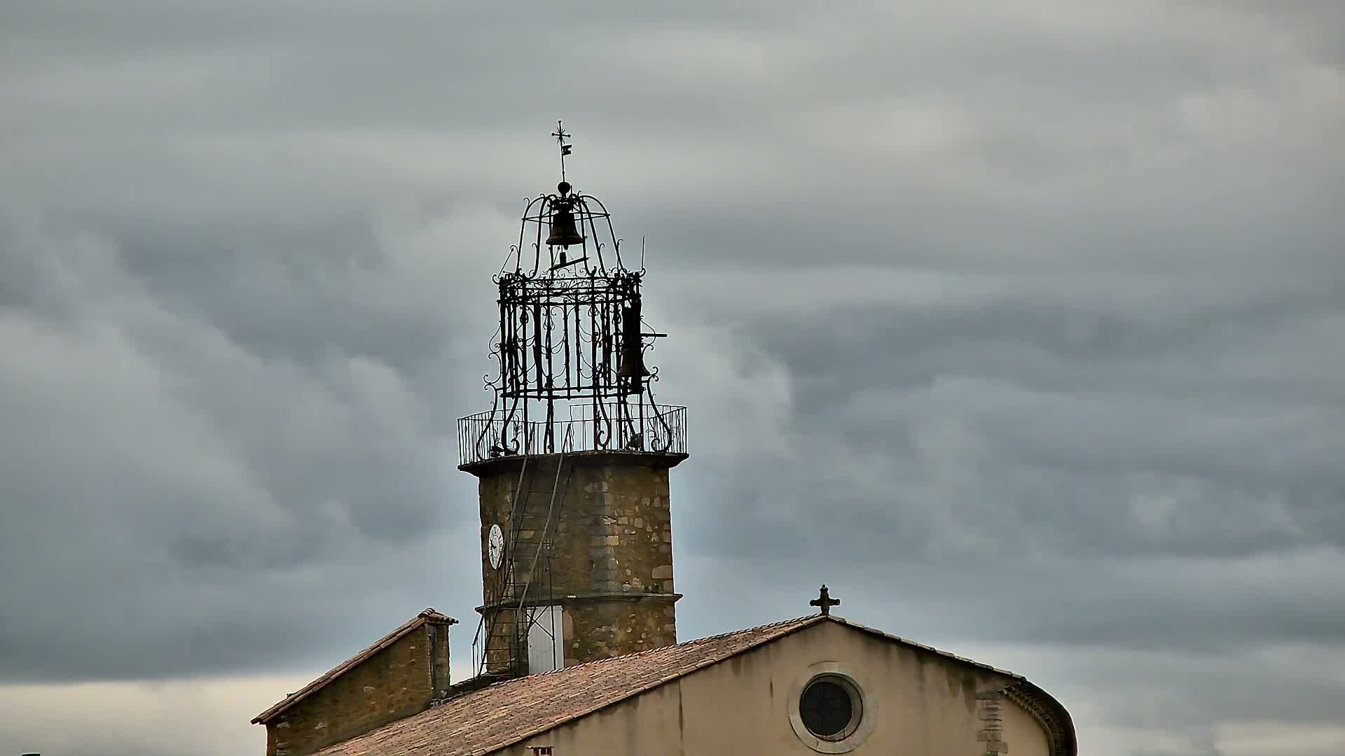 A prominent stone tower topped with an elaborate wrought-iron bell frame and parts of an adjacent church building with a tiled roof are framed against a foreboding, heavily overcast sky.