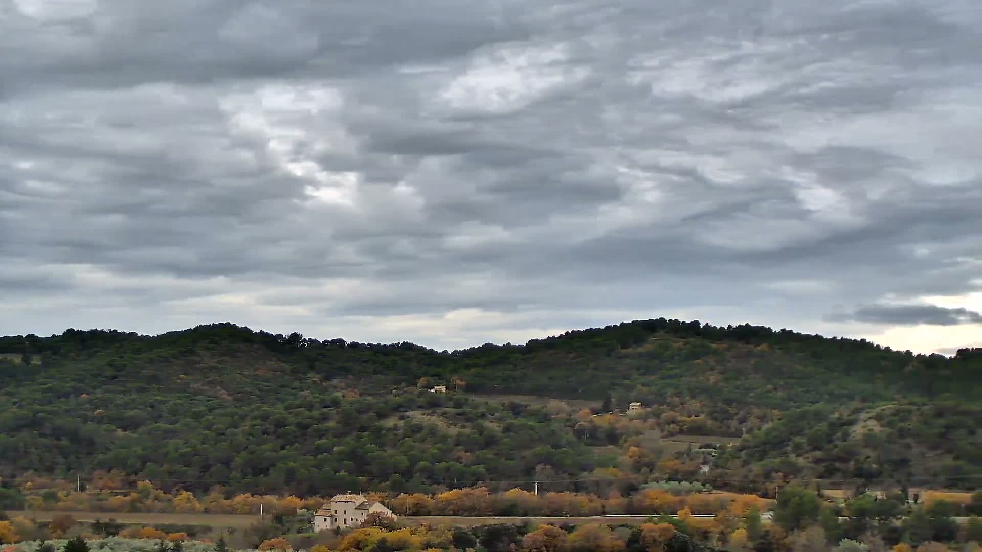 Under a heavily overcast and grey sky, a rural landscape unfolds with rolling hills covered in a mix of green and vibrant autumn foliage, dotted with a few distant houses.