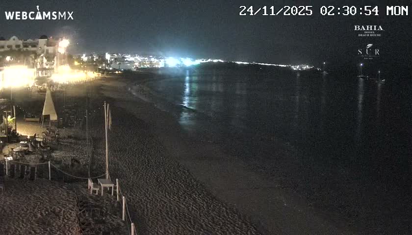 A dark sandy beach, lined with illuminated seating and buildings, extends alongside a calm ocean reflecting distant city lights and featuring several boats, all under a clear night sky.