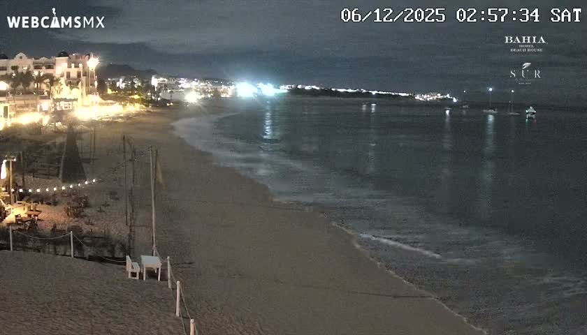 A serene night scene captures a resort beach with tables and string lights on the sand, gentle waves meeting the shore, and several boats anchored in the calm ocean under a clear, dark sky, with distant city lights illuminating the horizon.